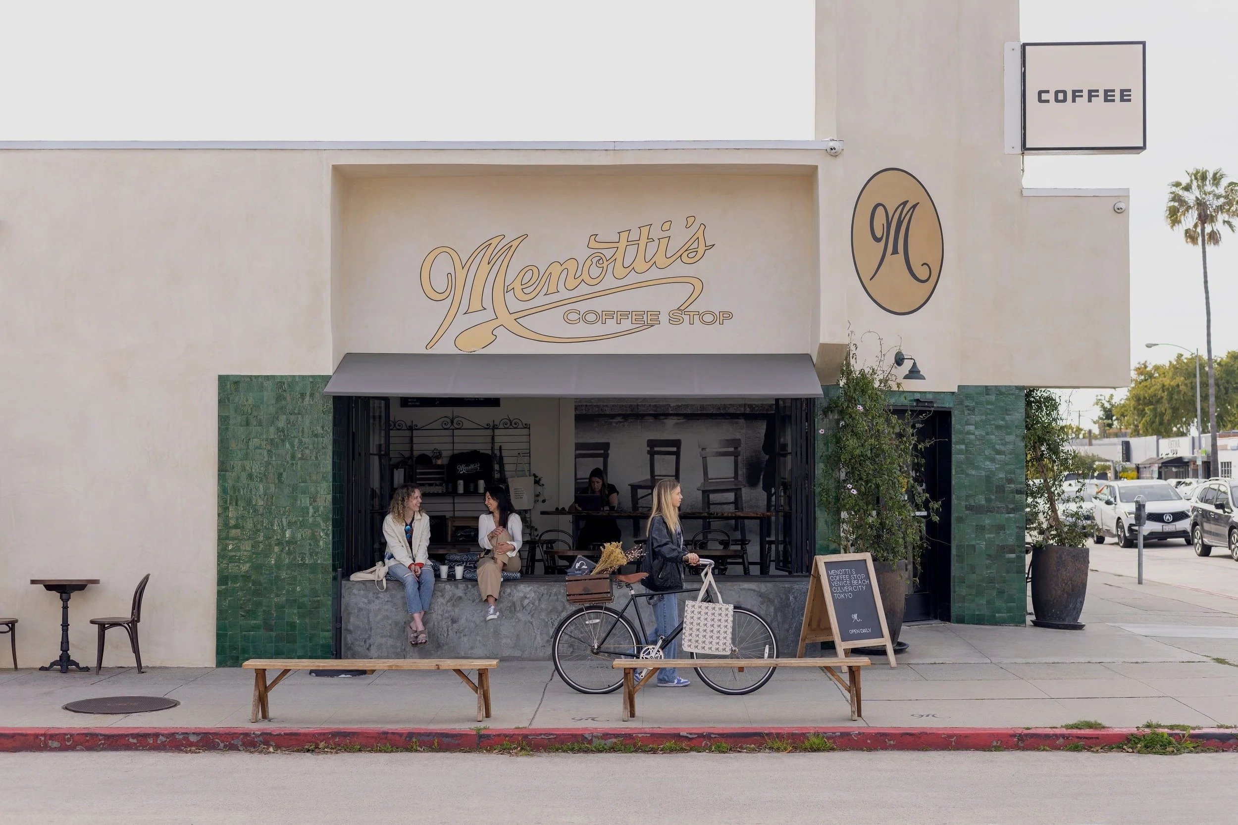 Exterior view of Menotti's Coffee Stop with outdoor seating, customers sitting and chatting, and a person riding a bicycle in front of the shop. Signage includes the shop's name and the word 'Coffee'.
