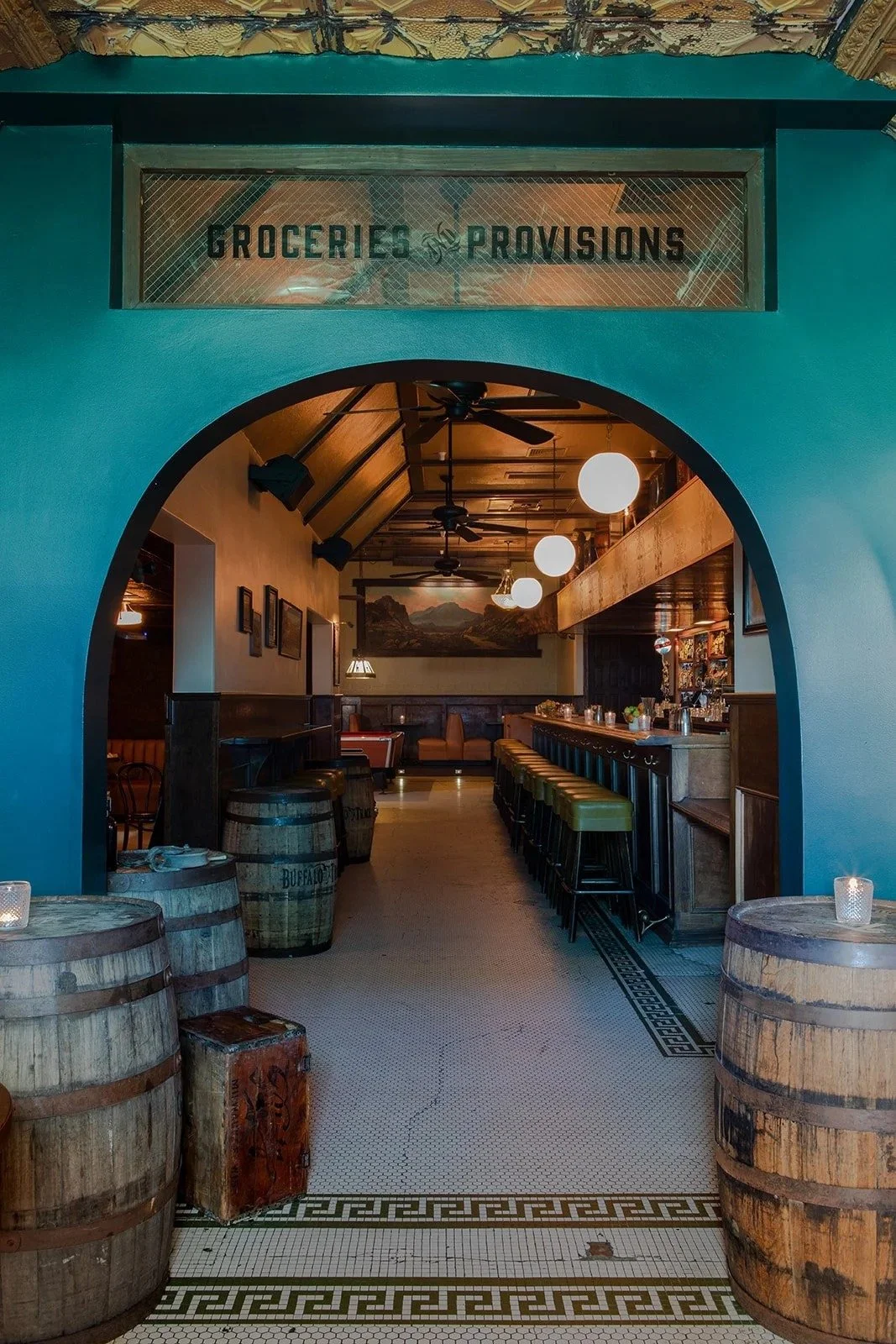 Interior of a restaurant with wooden barrels, a bar counter with green stools, warm lighting, and wall art, seen through an arched entryway with a sign above that reads 'Groceries & Provisions'.