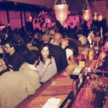 Crowded bar with people socializing and drinking, warm lighting, and bar counter with glasses and bottles.