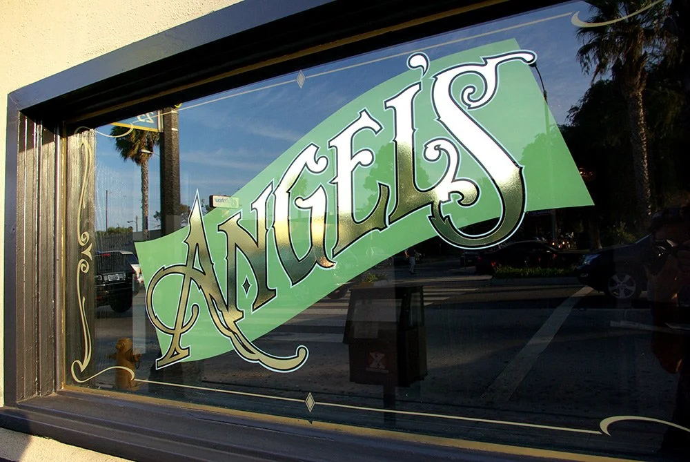 A window with a sign reading "Angels" in decorative white and green lettering, reflecting palm trees and parked cars outside.