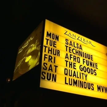 Illuminated marquee sign with schedule and event names, including Salsa Technique, Afro Funk, The Goods, Quality, and Luminous Mvv, displayed in black text on yellow background.