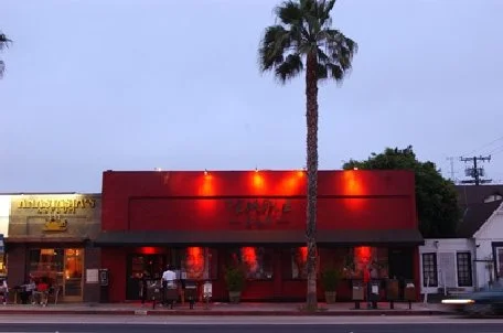 A red building with neon lights, two palm trees, and people standing outside during dusk.