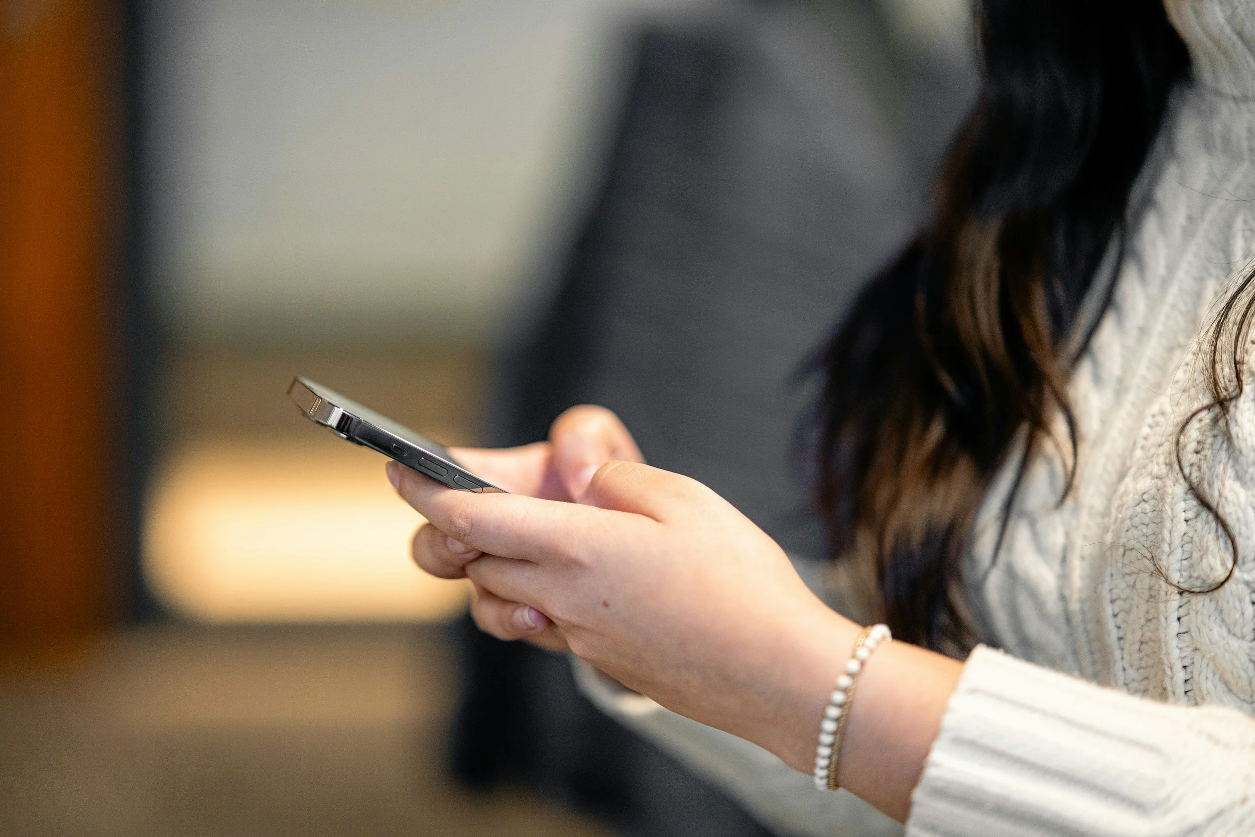 A person is holding a smartphone in their hand, with their thumb on the screen. The person has long, dark, wavy hair and is wearing a cream-colored sweater. There is a white beaded bracelet on their wrist.
