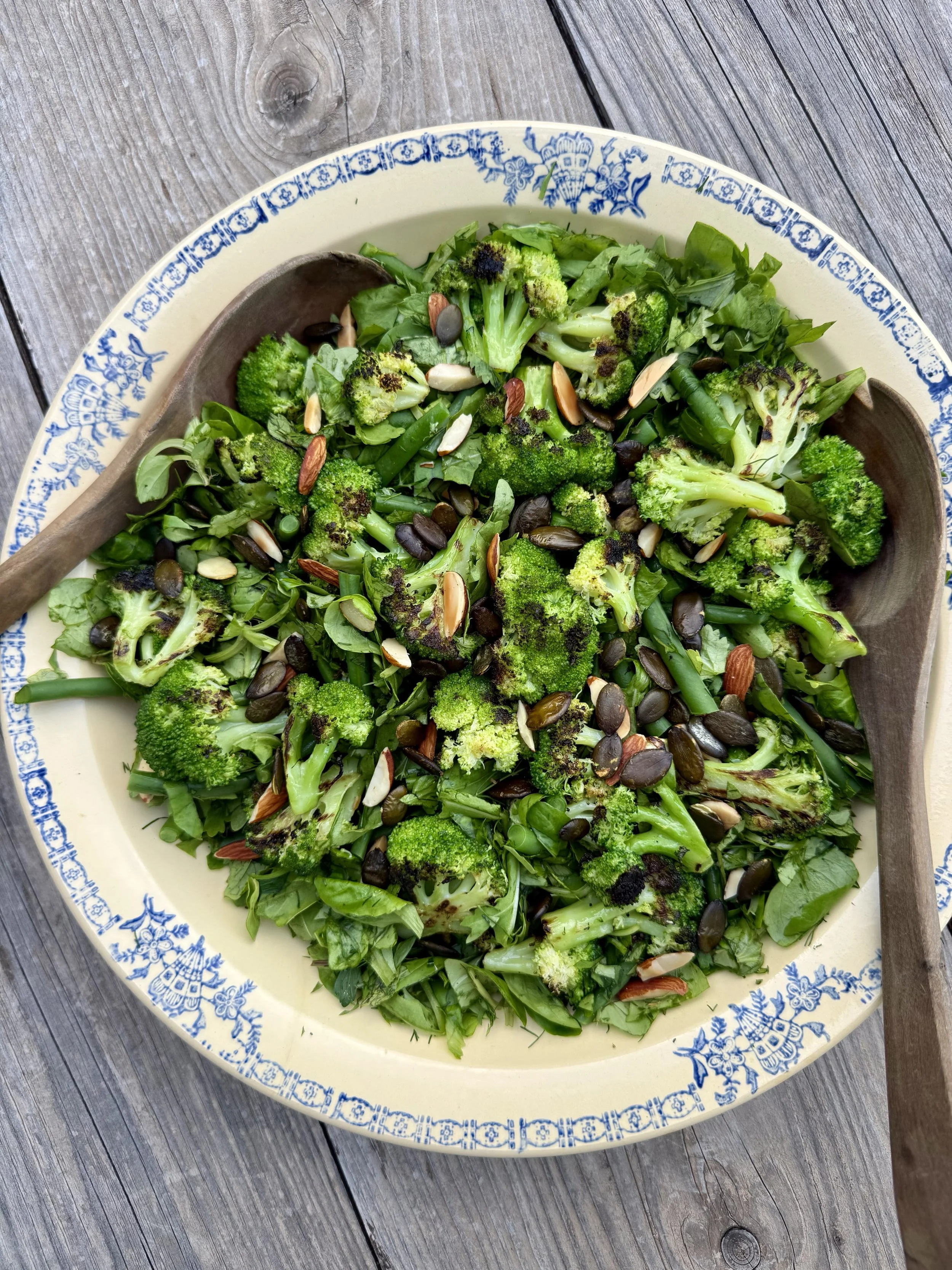 A decorative bowl of green salad with broccoli, green beans, spinach, and sliced almonds on a wooden table.