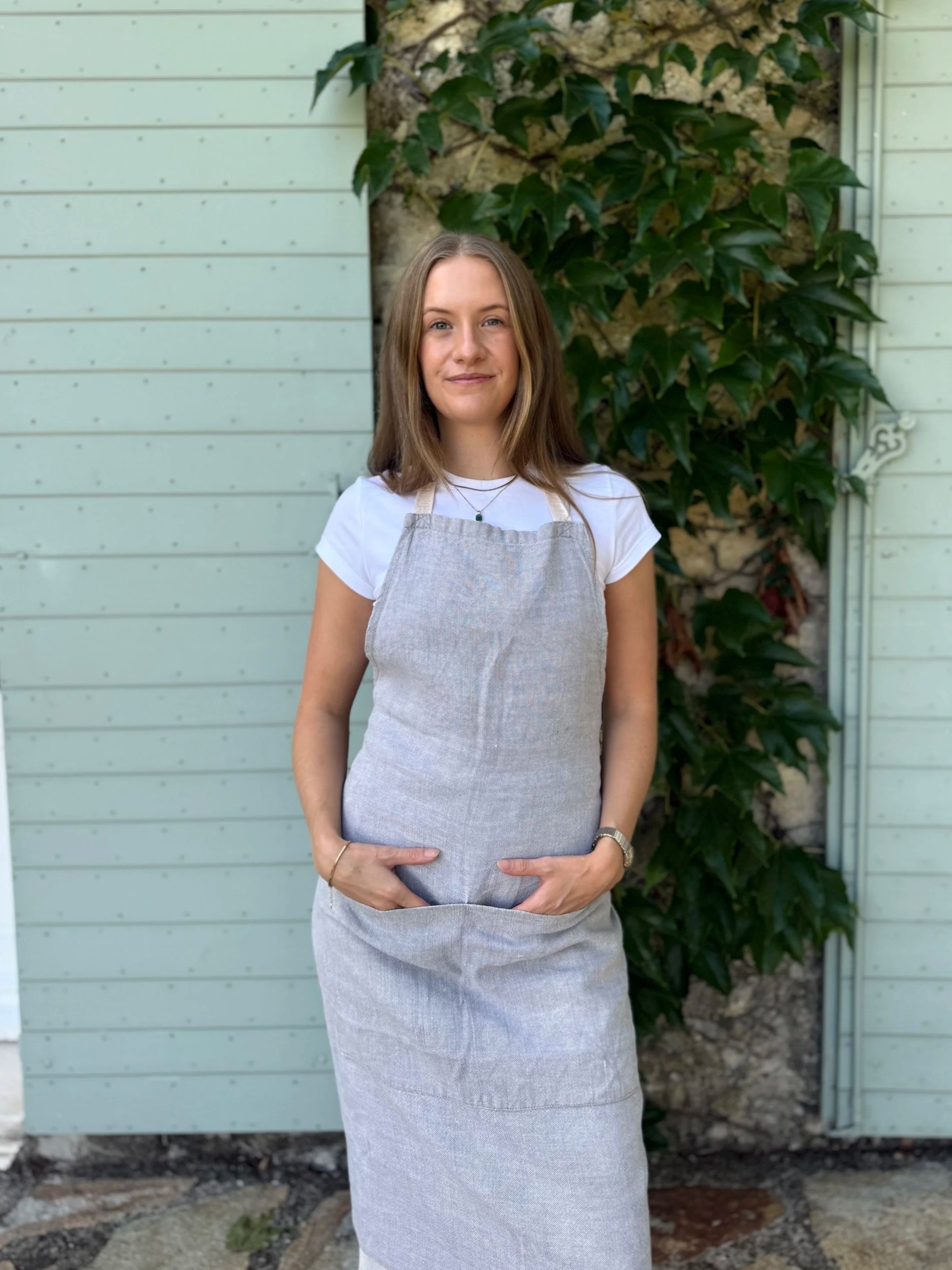 A young woman with shoulder-length blonde hair, wearing a white t-shirt and a light blue apron, standing outdoors in front of a light green wooden wall and a vine-covered stone wall, smiling softly.
