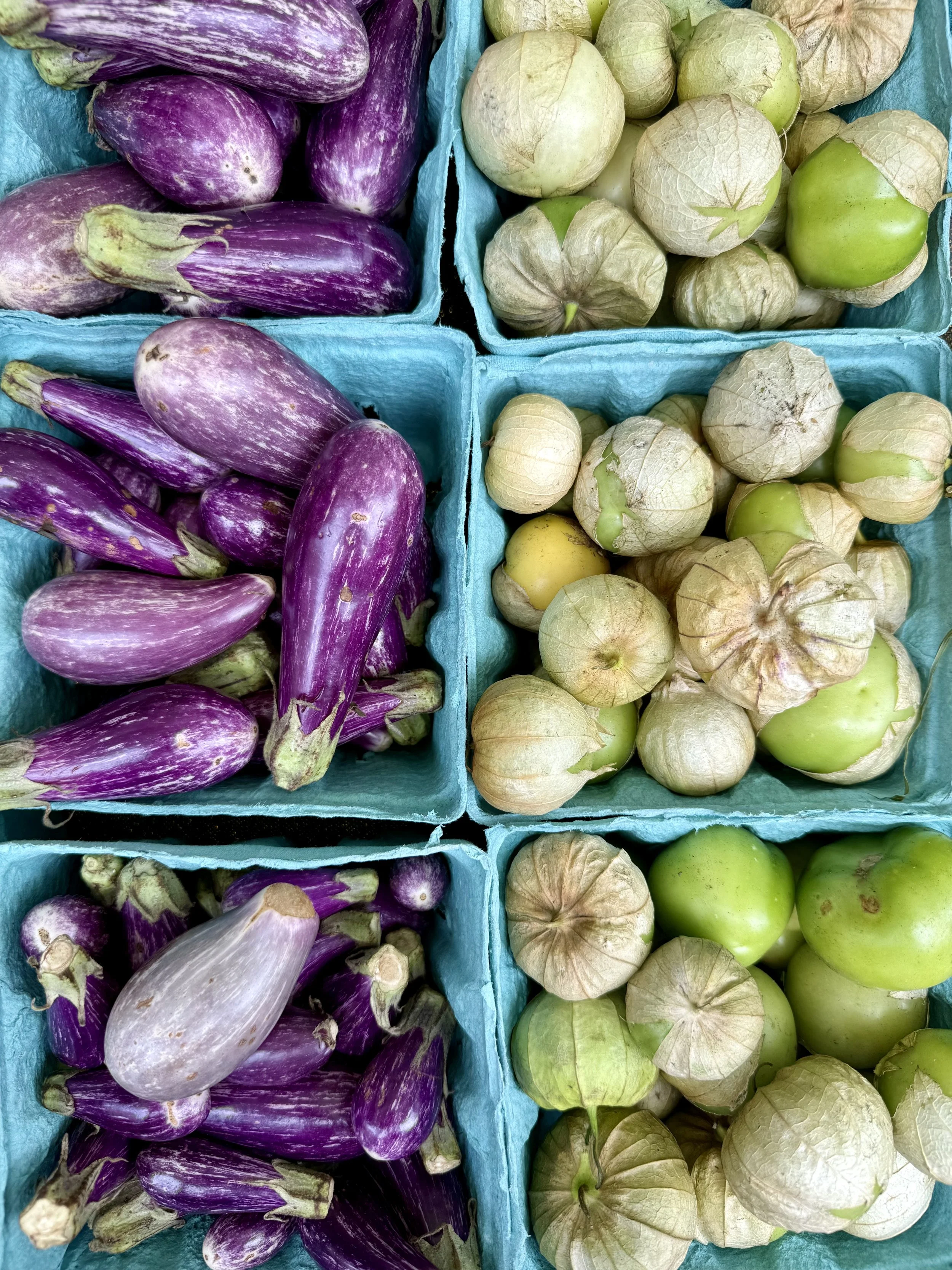 Four cardboard baskets filled with fresh vegetables: purple eggplants in the top left, green tomatillos in the top right, more purple eggplants in the bottom left, and green tomatillos in the bottom right.