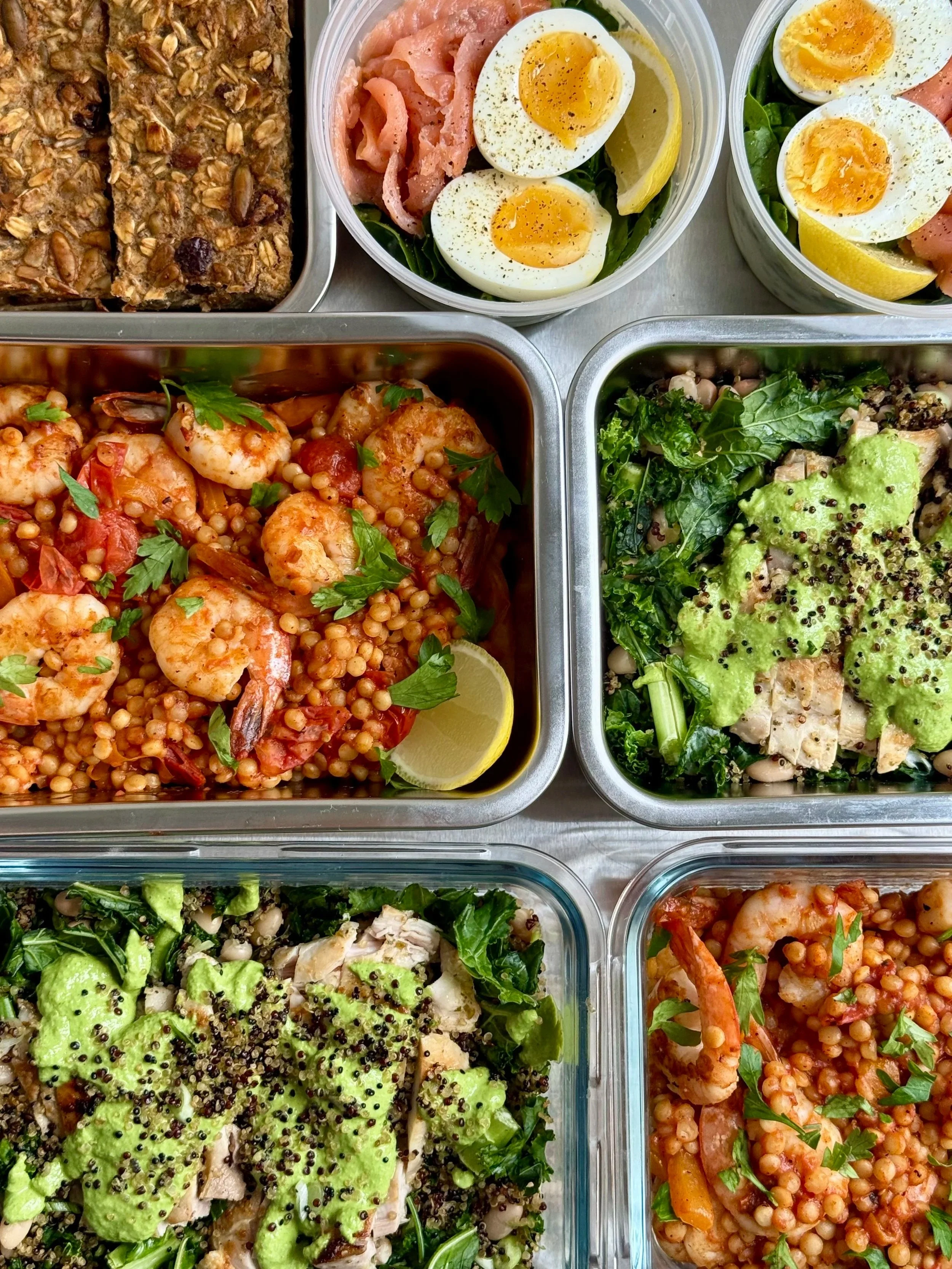 A variety of salads and dishes in metal containers including a shrimp dish with tomato and herbs, a roasted chicken salad with greens and mustard seeds, a green salad with avocado and black seeds, and two containers of boiled eggs with lemon wedges and herbs.
