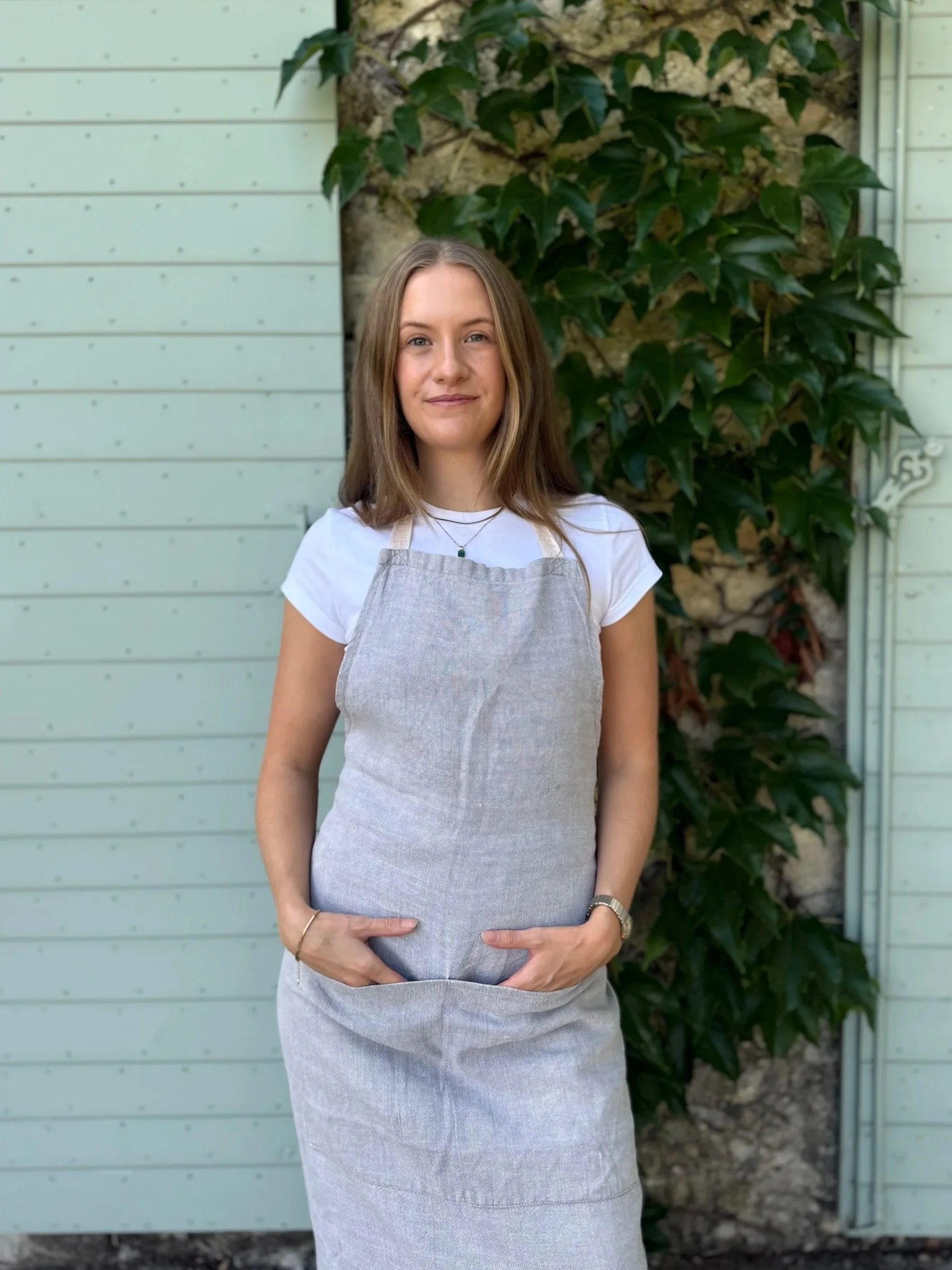 A young woman with shoulder-length brown hair, wearing a white t-shirt, a grey apron, and a necklace, standing outdoors in front of a light-colored wooden structure and green leafy plants, smiling and holding her hands on her stomach.