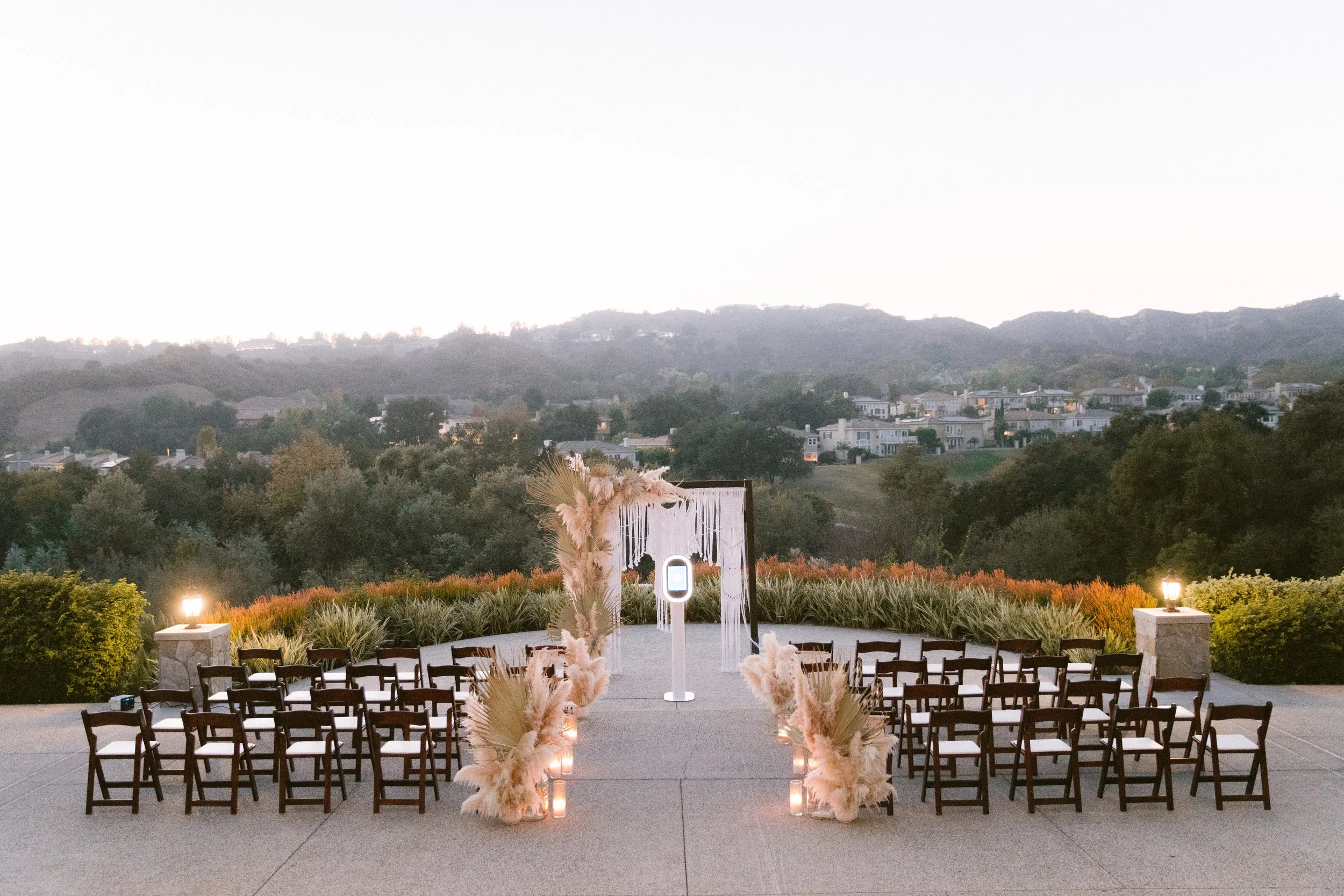 Outdoor wedding ceremony setup with chairs facing an altar decorated with pampas grass and candles, overlooking hilly landscape with houses and trees.