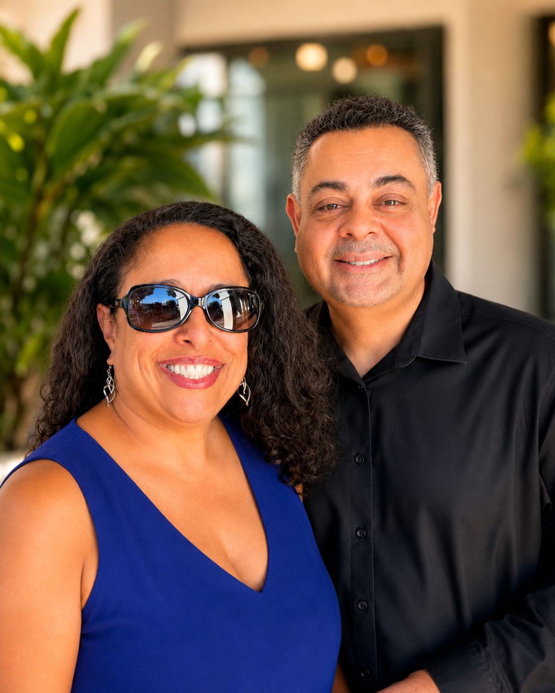 A smiling woman in a blue sleeveless dress wearing sunglasses and earrings standing next to a smiling man in a black shirt outdoors with greenery in the background.