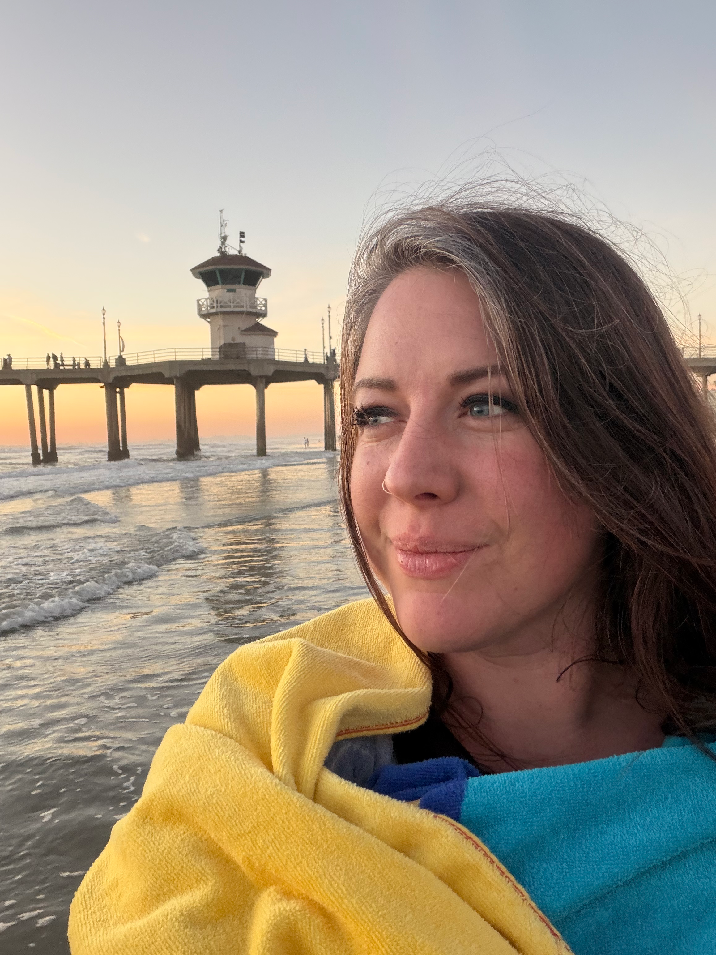 A woman with brown hair and blue eyes at the beach during sunset, smiling slightly, wrapped in a multicolored towel, with a pier and ocean in the background.