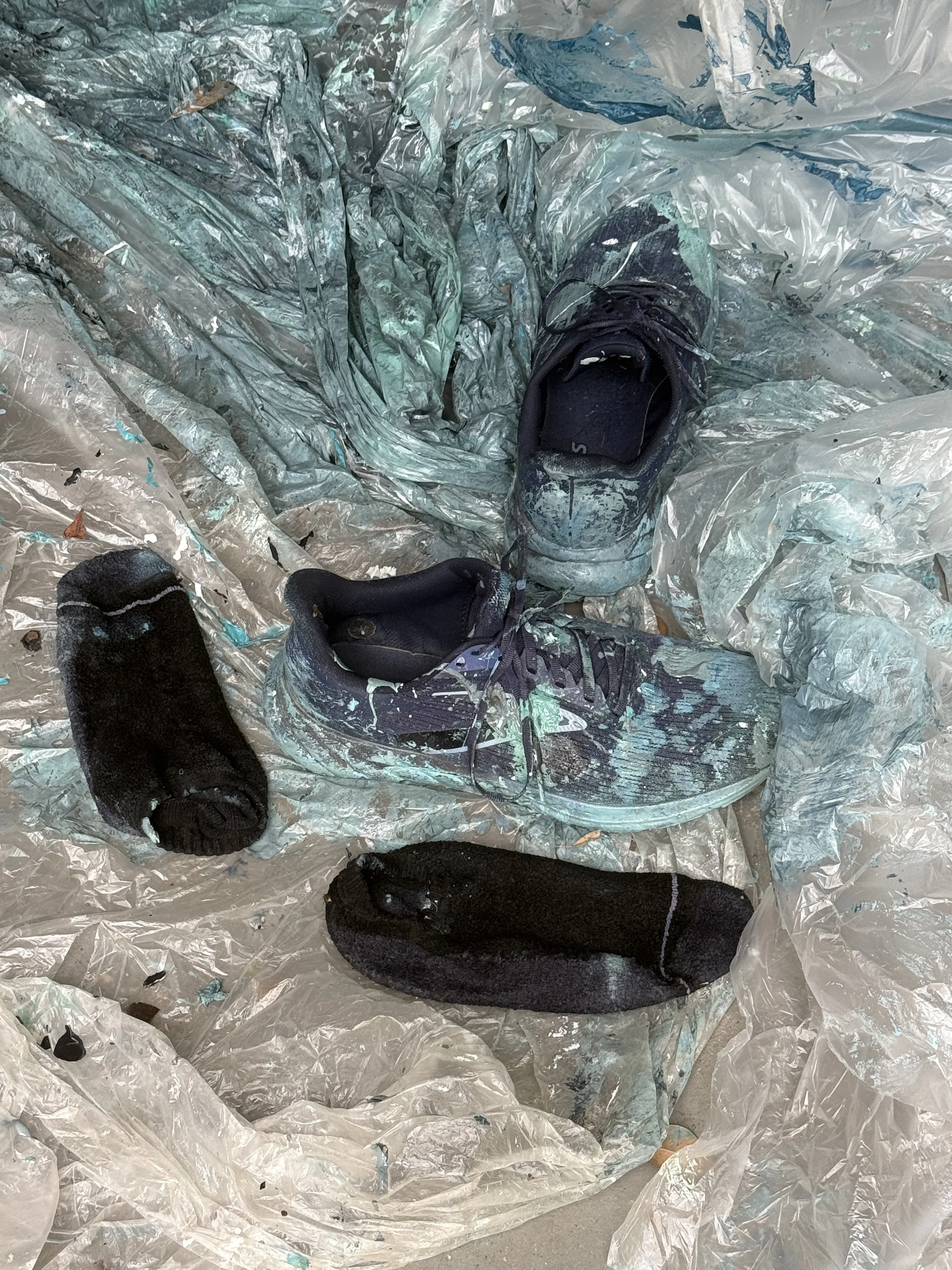 A pair of black running shoes with gray and multicolored details placed on top of crumpled plastic sheets, with two black socks nearby.