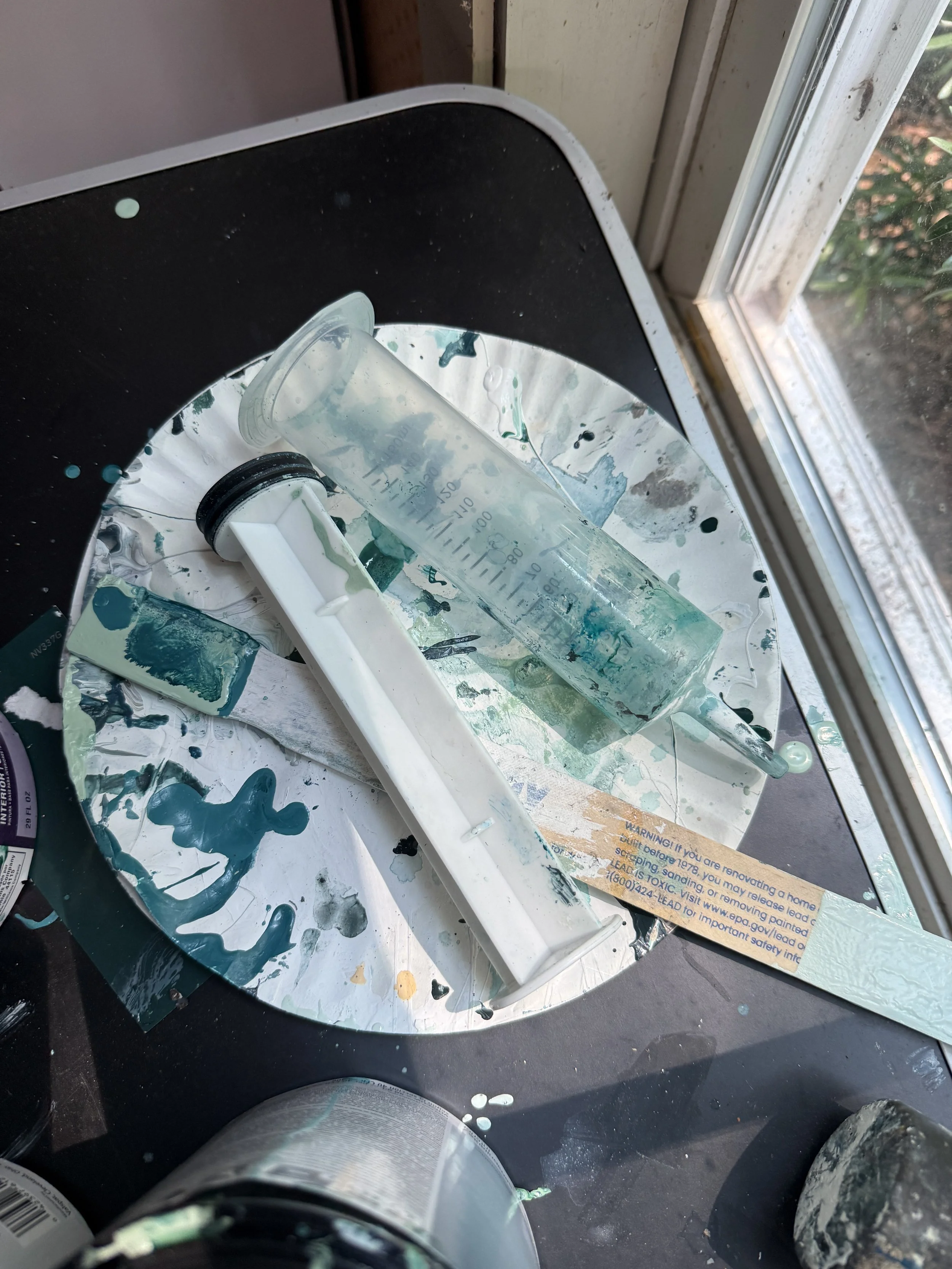 The image shows a messy artist's workspace near a window. There are paint splatters on the surface, a plastic cup with paint remnants, a palette with mixed paint colors, and a paint tube. The workspace appears to be used for painting or creating art.