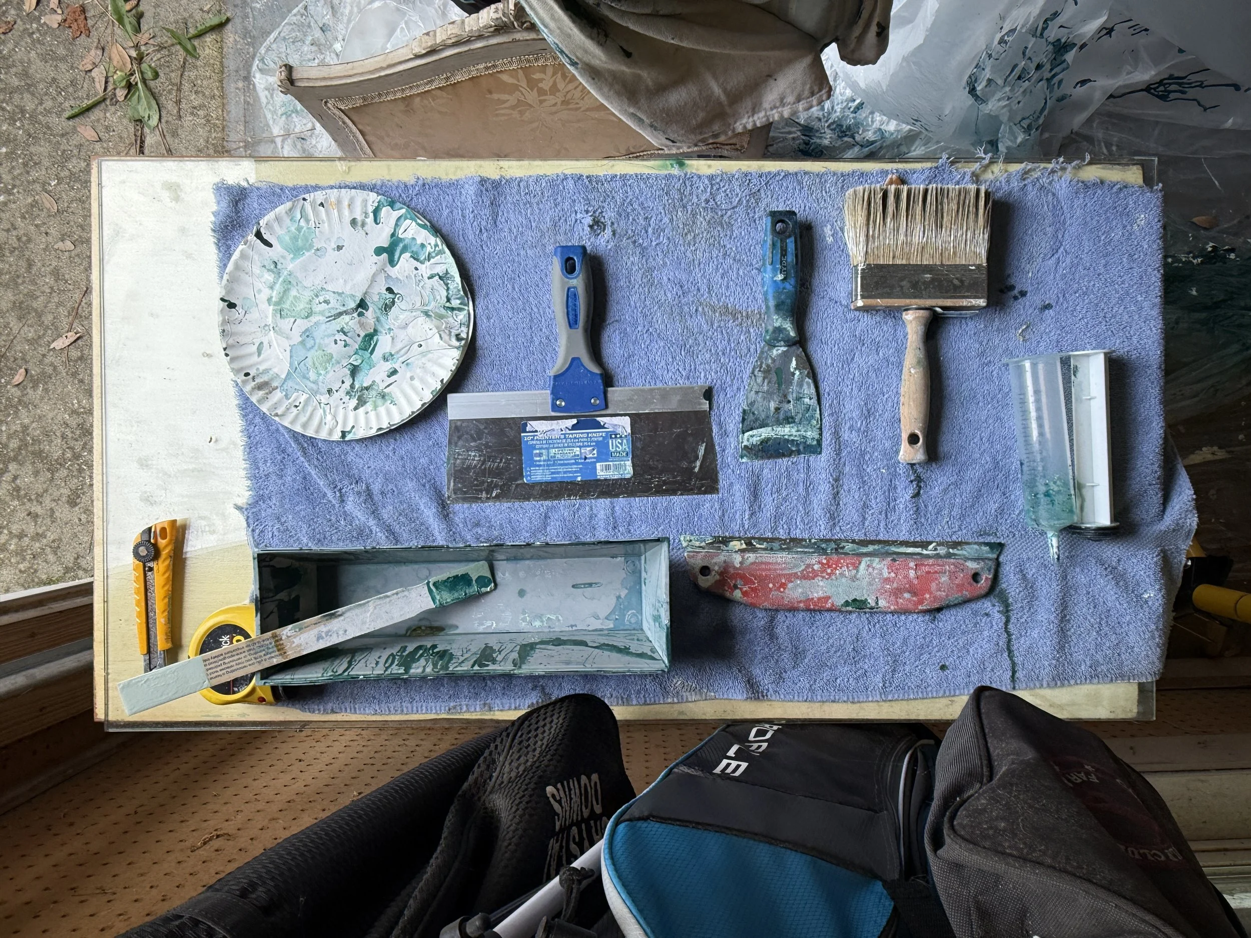 A workbench covered with a blue towel, with art supplies including a painted paper plate, a putty knife, a paintbrush, a paint roller, and a container of white paint. Part of a person's leg in black pants is visible at the bottom of the image.
