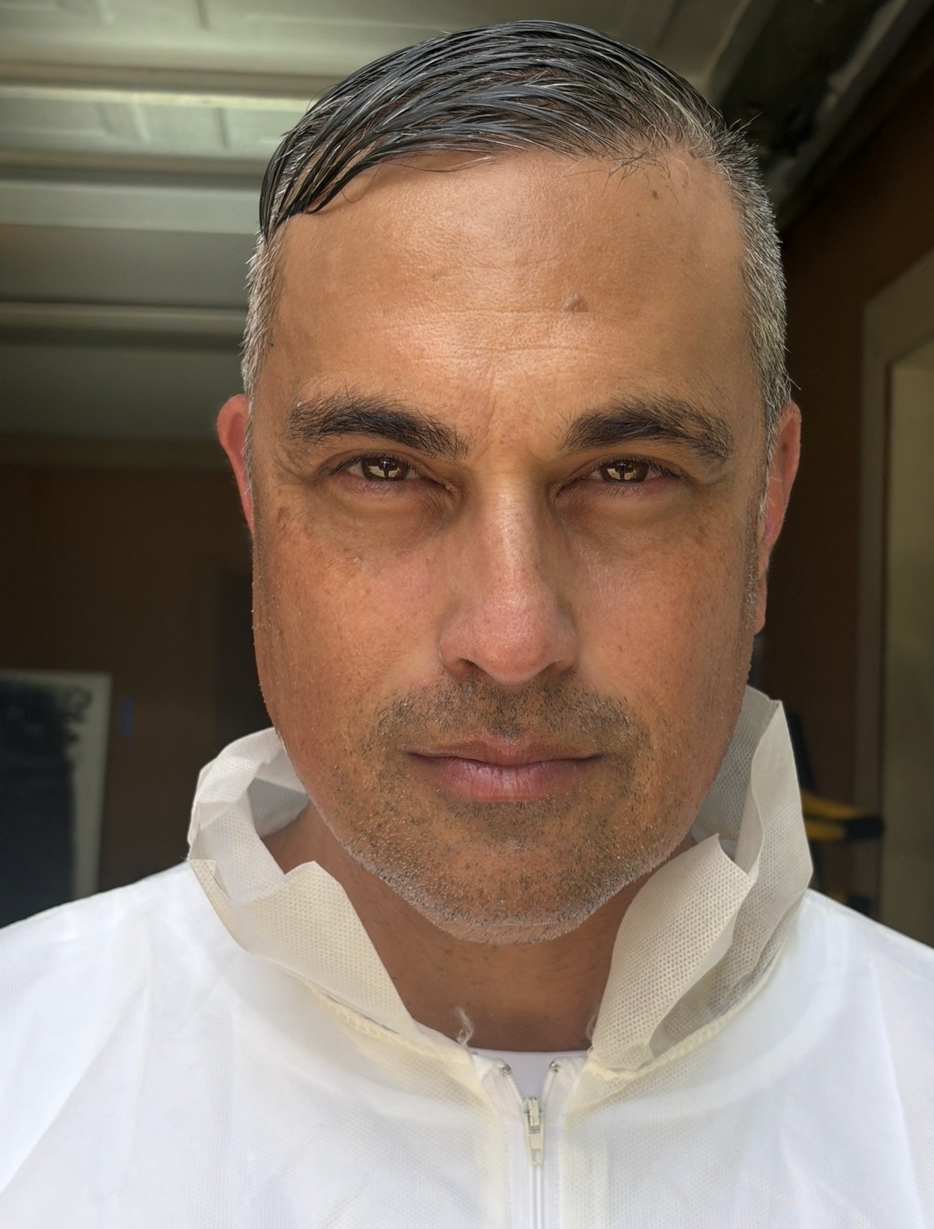 A man with short, dark, and slightly graying hair, wearing a white protective medical gown and mask pulled down, standing indoors with a neutral background.