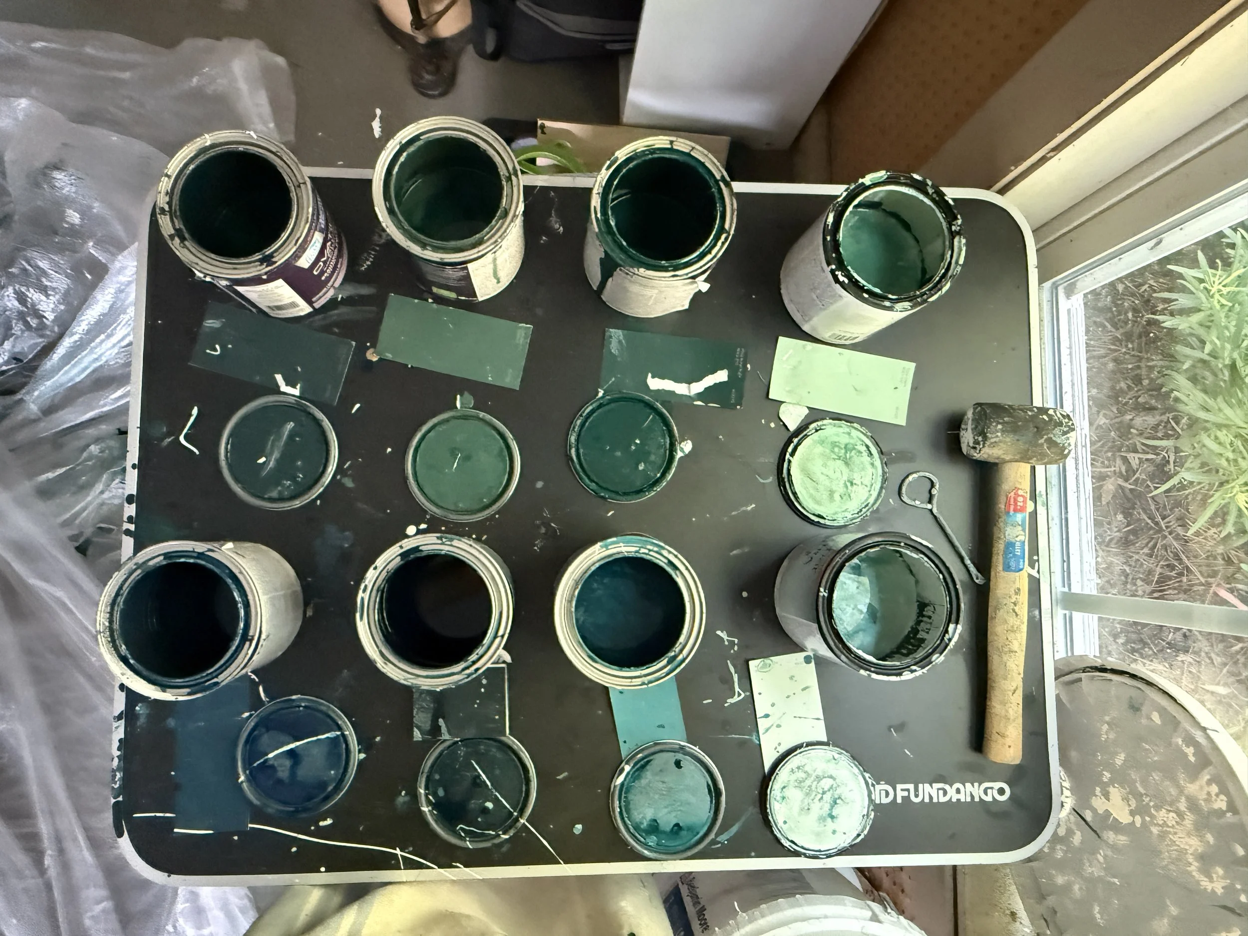 Open cans of green paint on a black surface, with paint brushes and a small hammer nearby, near a window with trees outside.