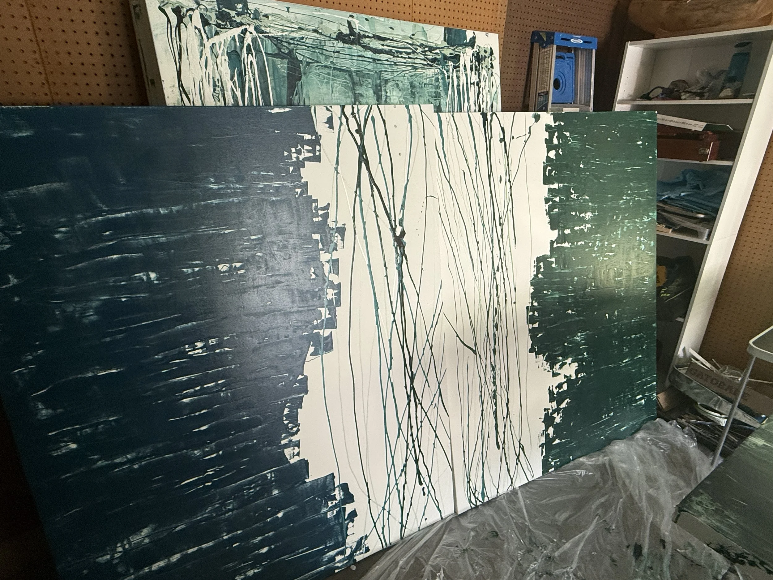 Abstract painting leaning against a pegboard wall, featuring black, white, green, and blue splashes and strokes with a dripping effect.