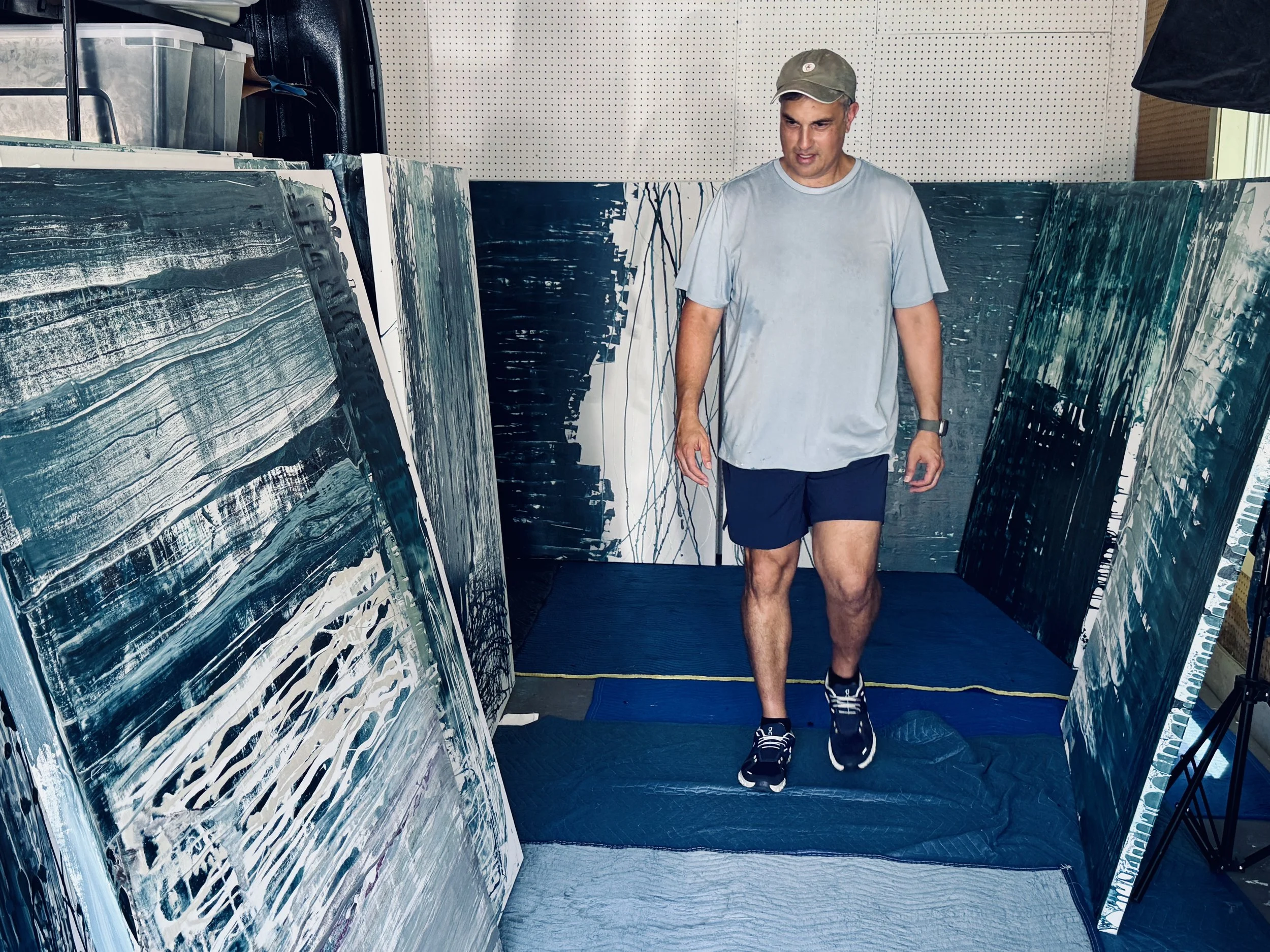A man looking at abstract paintings in an art gallery, wearing a light gray t-shirt, dark shorts, sneakers, and a baseball cap.