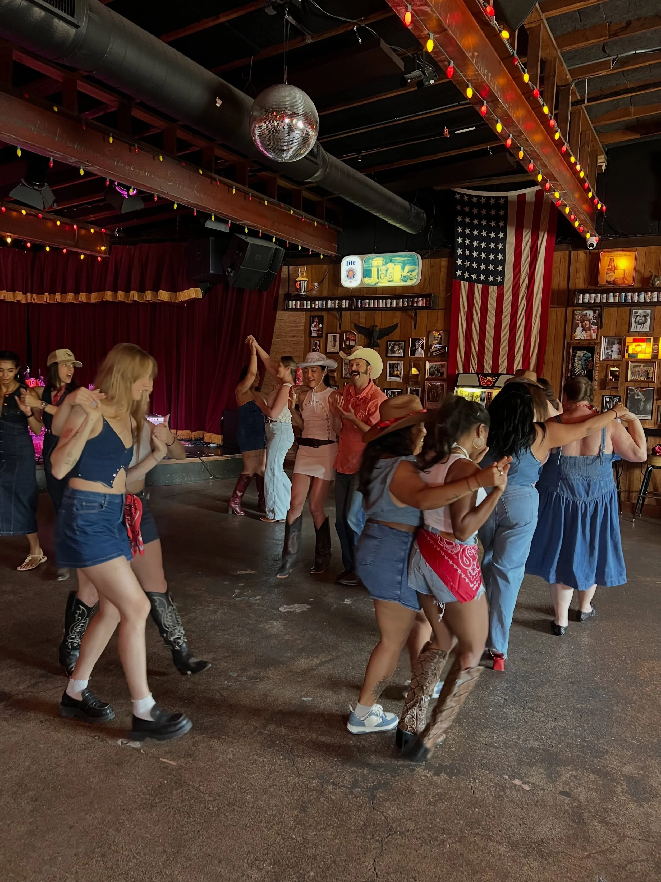People dancing at a country-themed bar or club with wooden walls, cowboy hats, and American flag decorations.