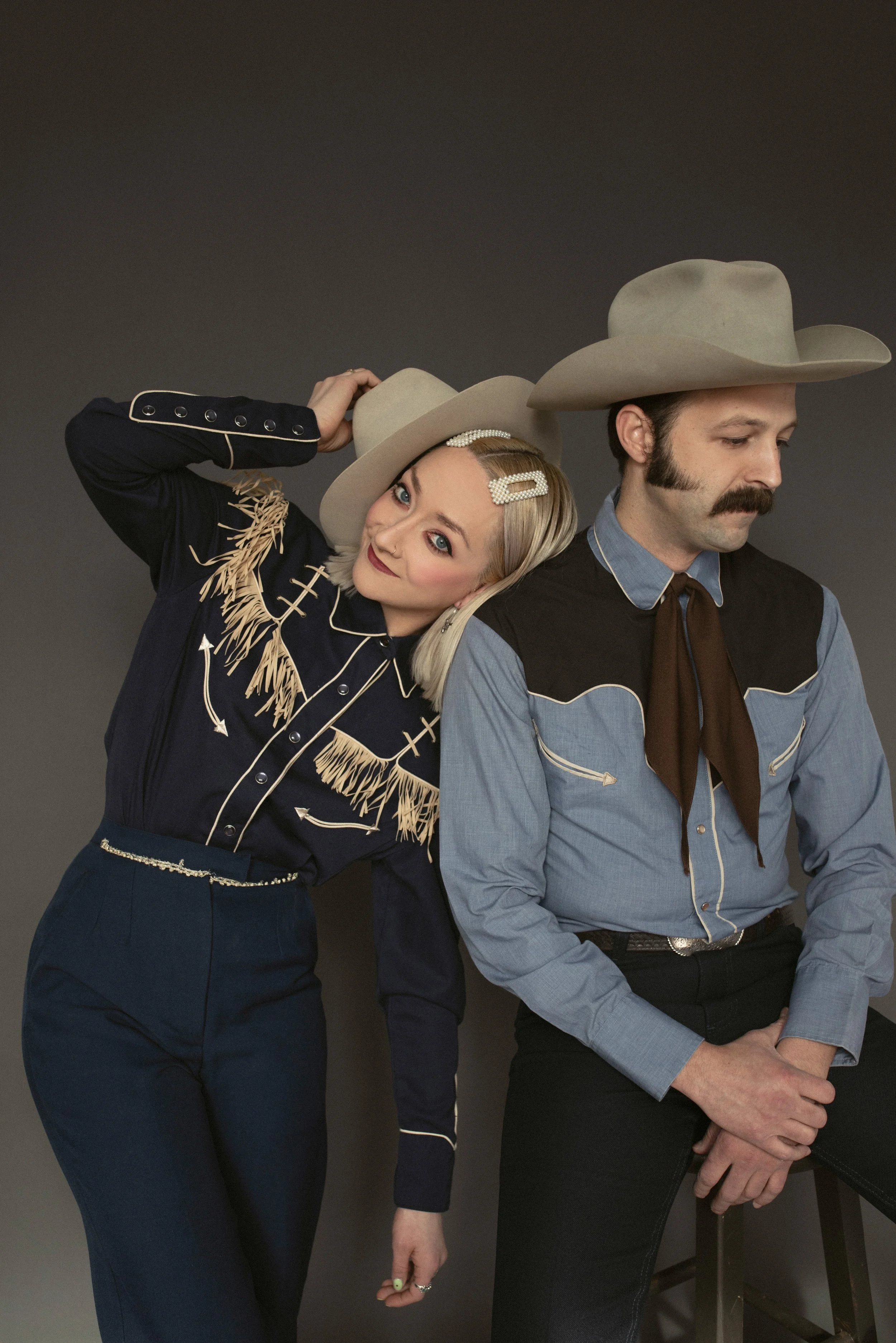 Two people dressed in western-themed attire, with the woman wearing a pearl-decorated hair clip and a fringed western shirt, and the man wearing a cowboy hat, blue western shirt, and brown neckerchief, pose against a dark background, with the woman holding her cowboy hat and leaning on the man's shoulder.