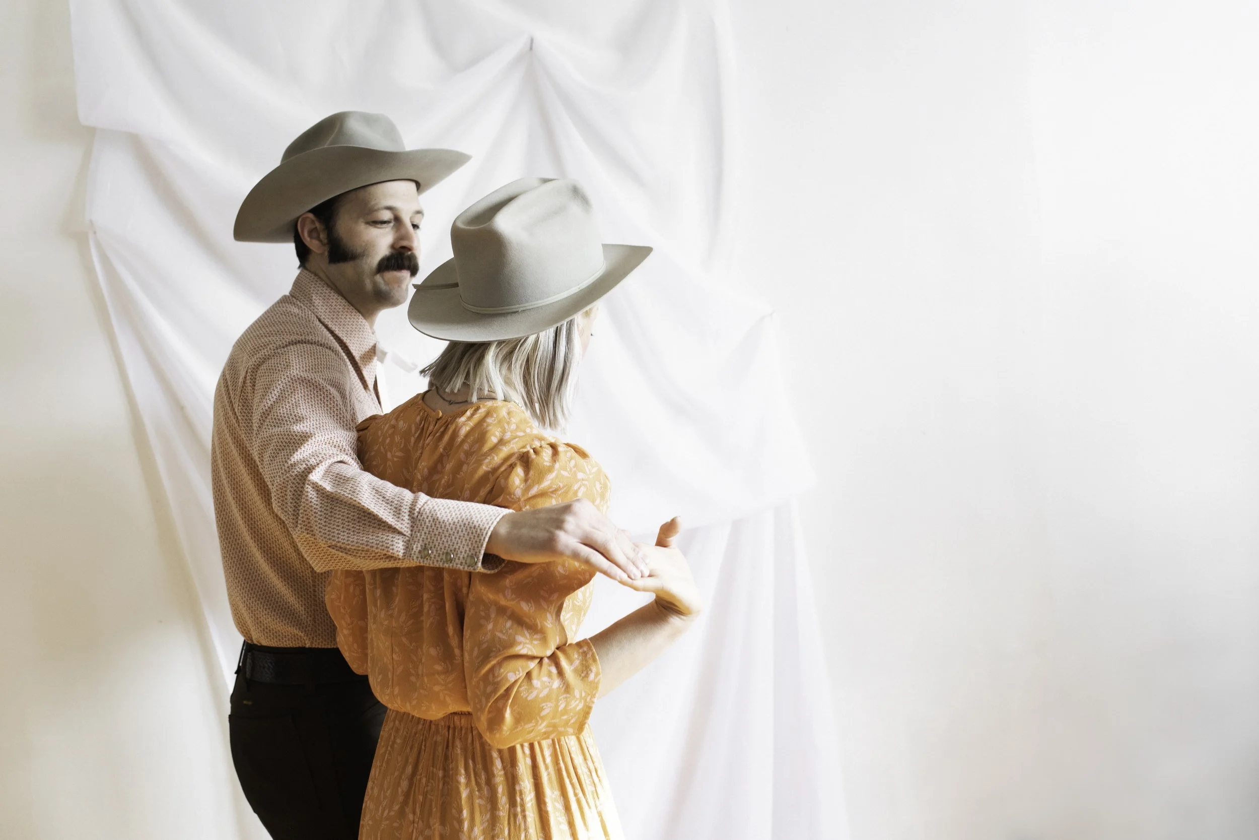 A man with a mustache wearing a cowboy hat, a patterned shirt, and black pants standing behind a woman in a long orange dress and a light-colored hat, both holding their hands out as if dancing or practicing a dance, with a white backdrop.