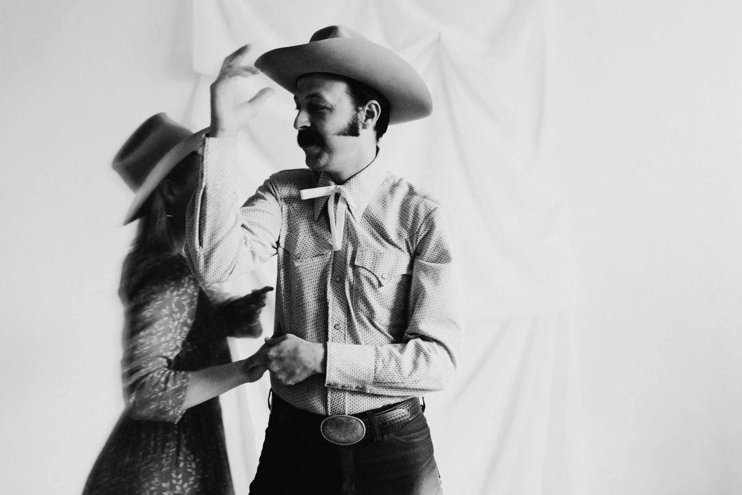 A black and white photo of a man wearing a cowboy hat, a patterned shirt, and a large belt buckle, as he dances with a person wearing a long-sleeved dress with leaf patterns, against a plain white background.