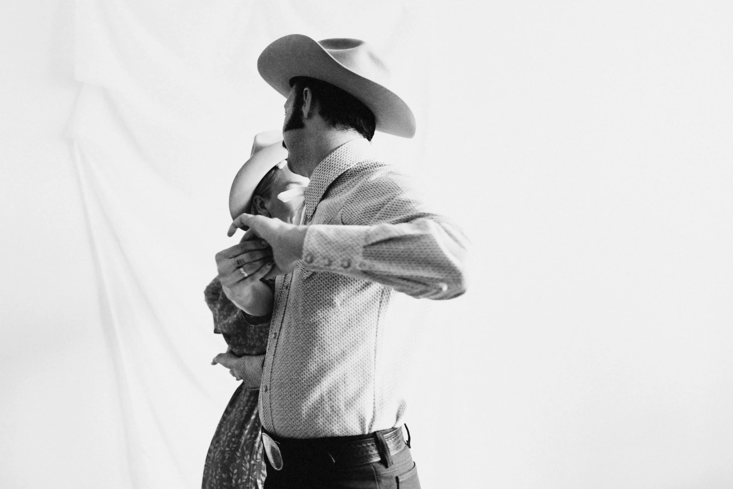 A black and white photo of a man wearing a cowboy hat, a polka dot shirt, and jeans, holding a young girl in a dress close to his face, both partially visible.