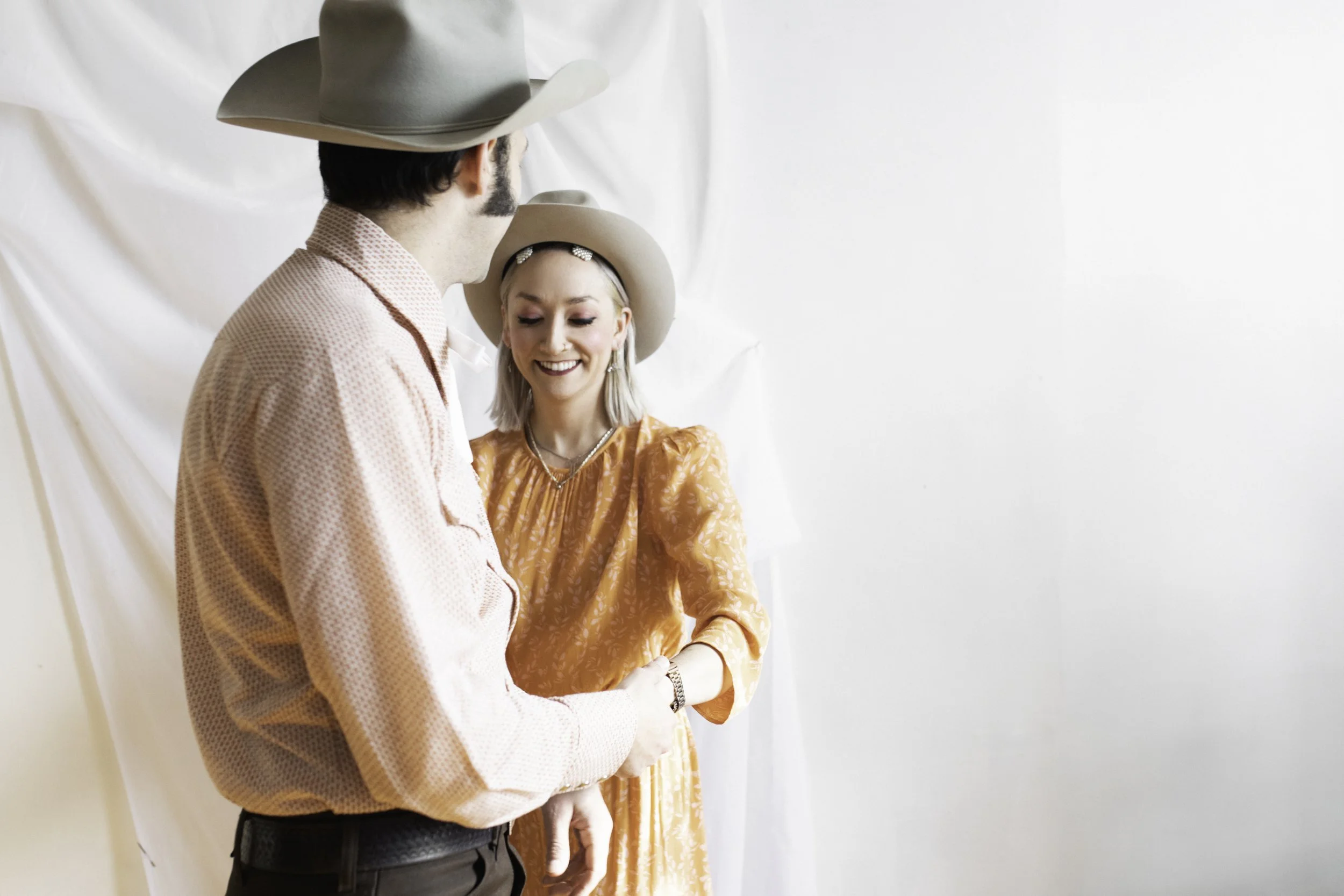A man and woman wearing cowboy hats smiling and interacting in front of a plain white background.