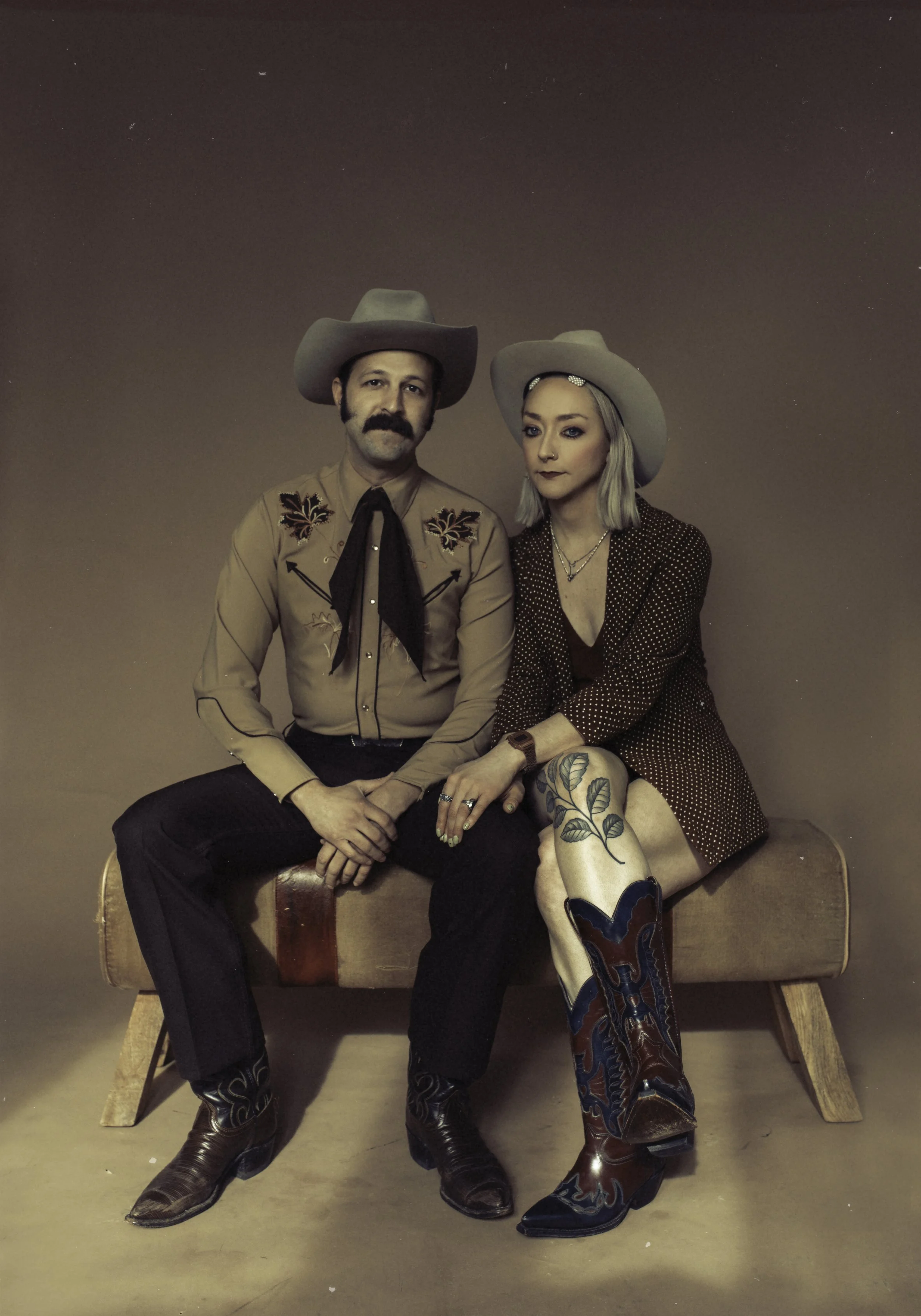 A man and woman dressed in Western-style clothing sitting on a wooden bench against a plain background. The man has a cowboy hat, mustache, Western shirt with embroidery, black pants, and cowboy boots. The woman has a cowboy hat, blonde hair, dark ma