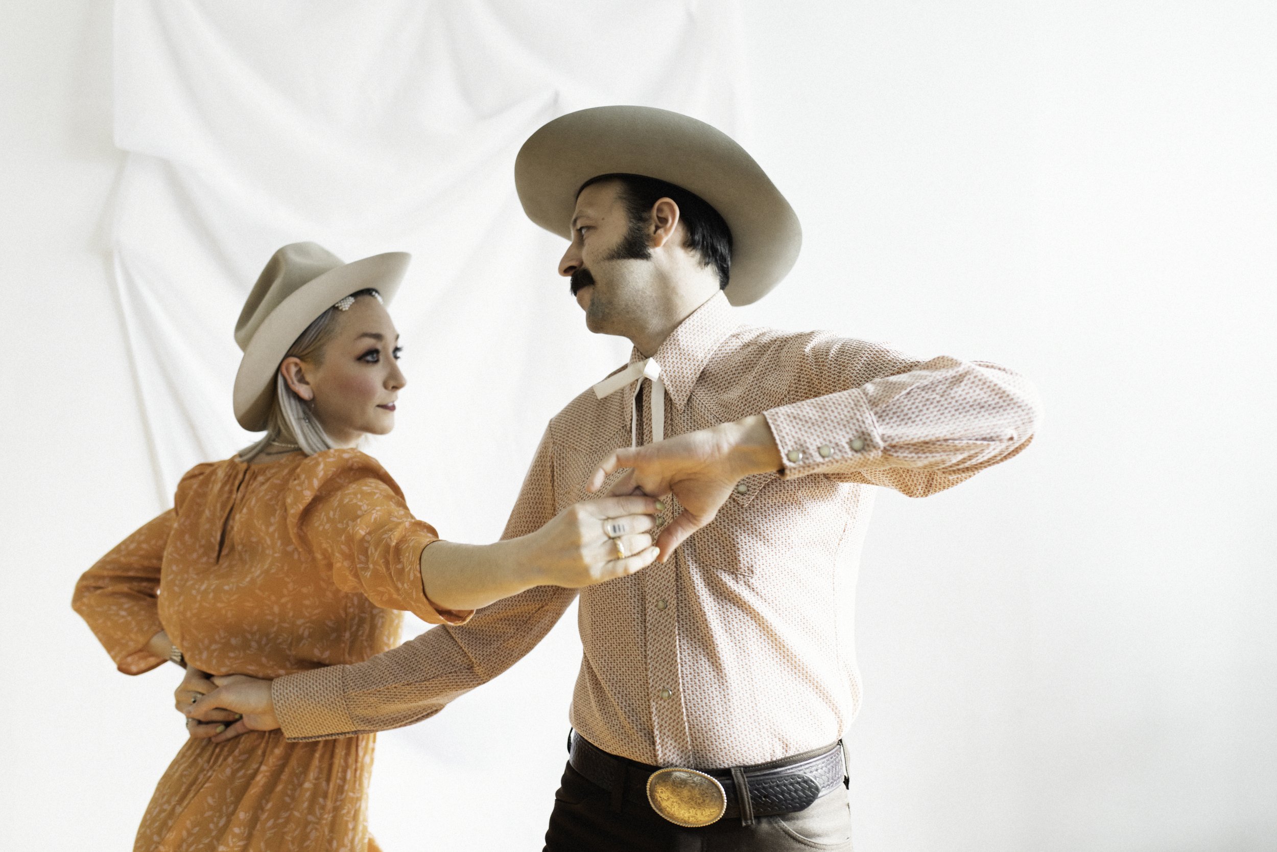 A man and woman dancing together against a white backdrop, wearing vintage Western-style clothing and hats, holding hands in a dance pose.
