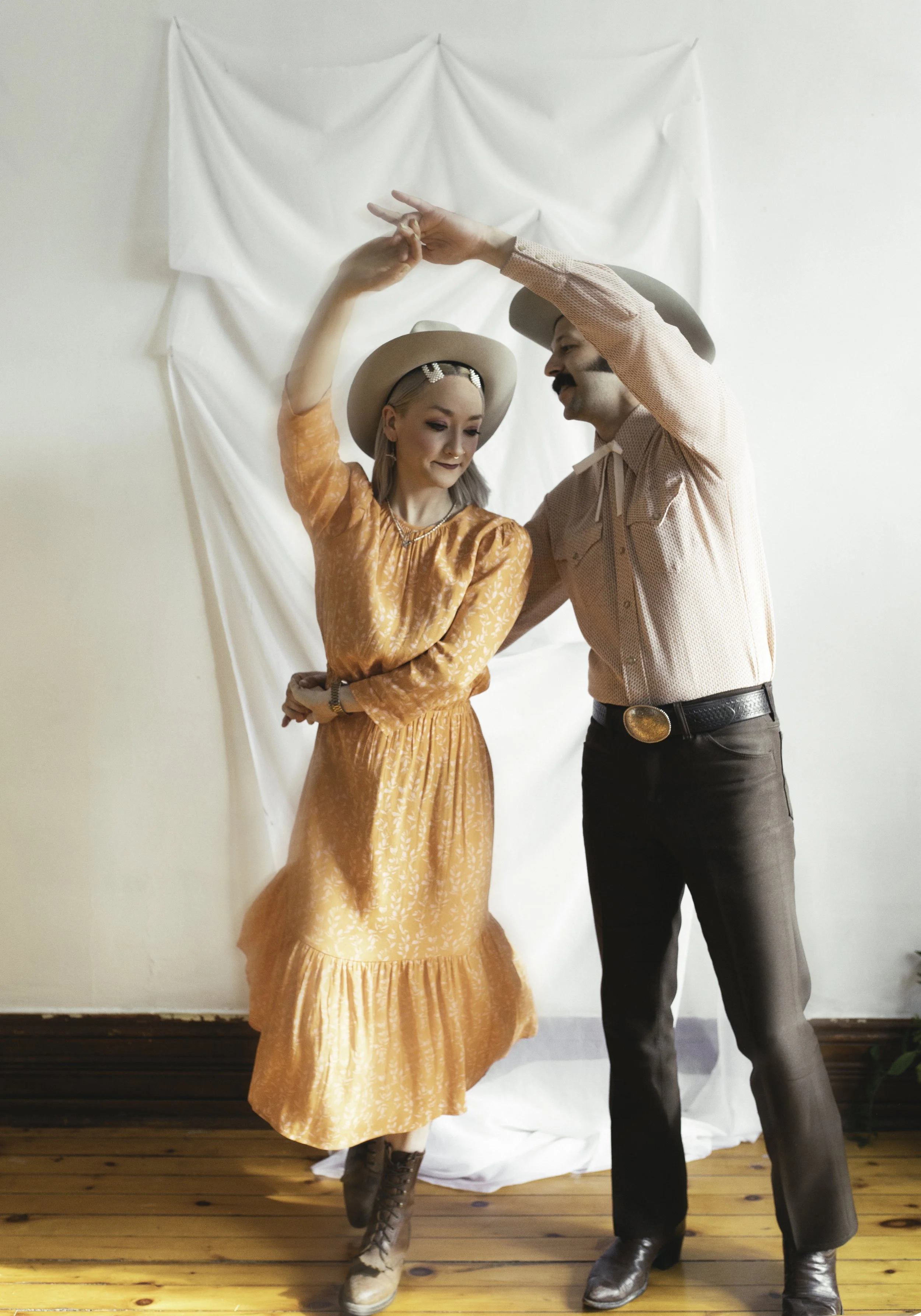 A couple in vintage western outfits dancing in front of a white backdrop on a wooden floor.