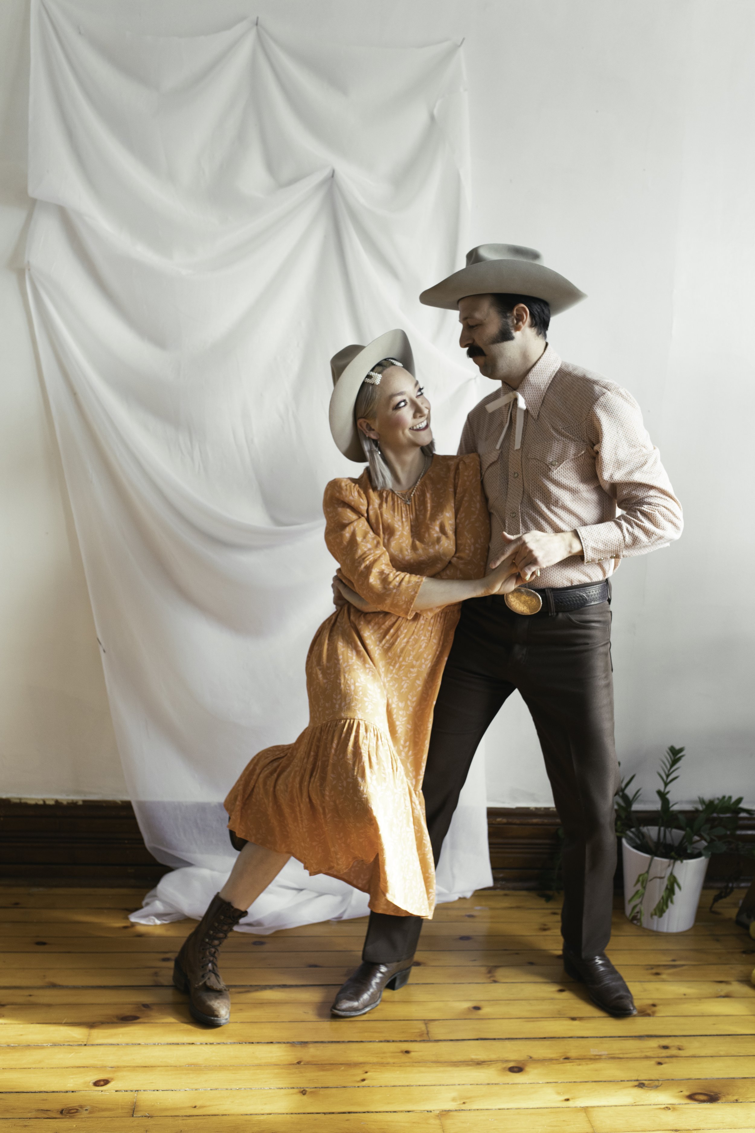 A man and woman dressed in cowboy outfits, standing close together and holding hands, in front of a white draped backdrop. The woman is smiling and looking at the man, who is looking down at her.