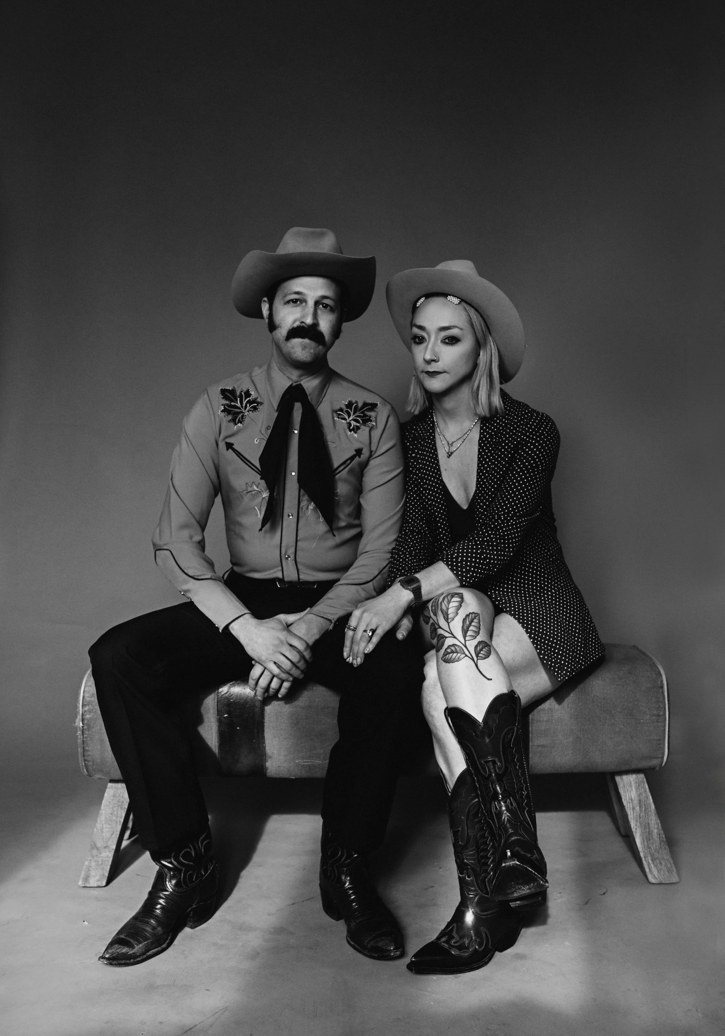 Black and white photo of a man and woman in cowboy outfits sitting on a bench. Both are wearing cowboy hats and cowboy boots, with the woman also wearing a polka dot jacket and the man a Western shirt with embroidered designs.