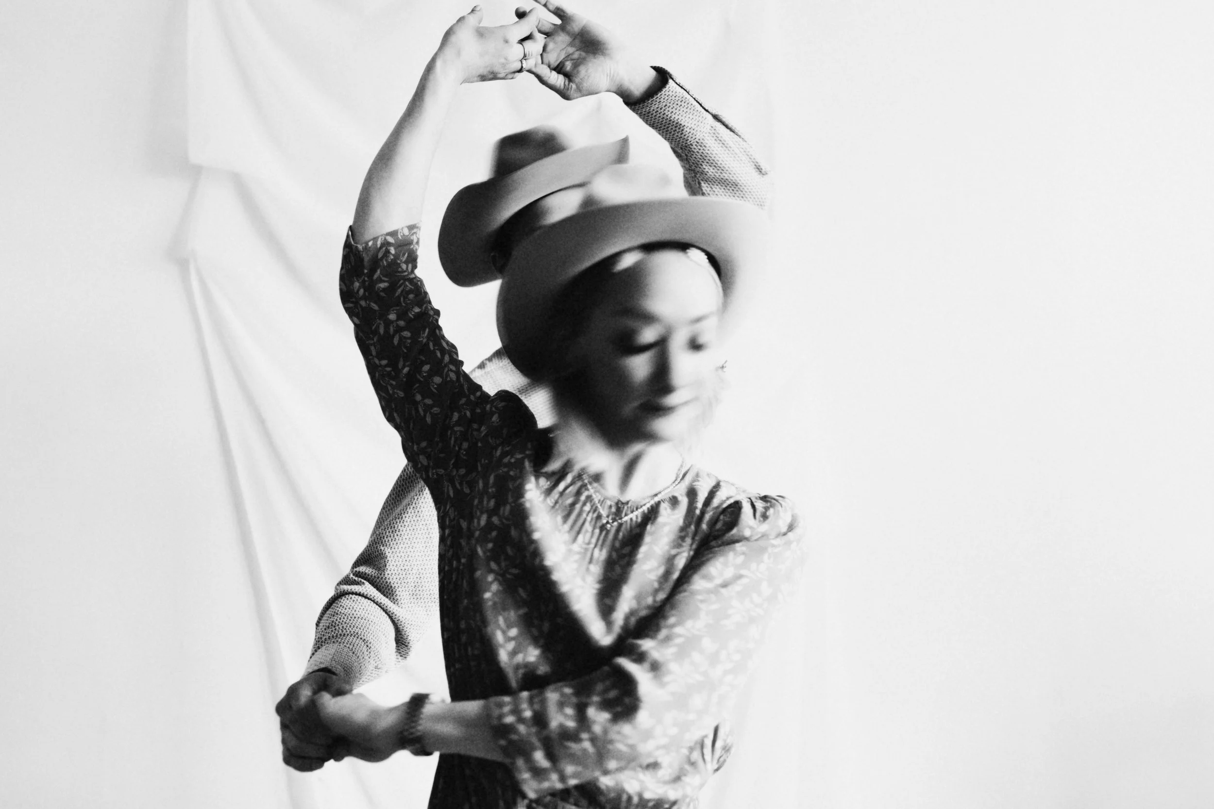 A woman with her eyes closed, wearing a floral patterned shirt and multiple rings, is balancing hats on her head while holding a hat in her hands, against a plain light background.