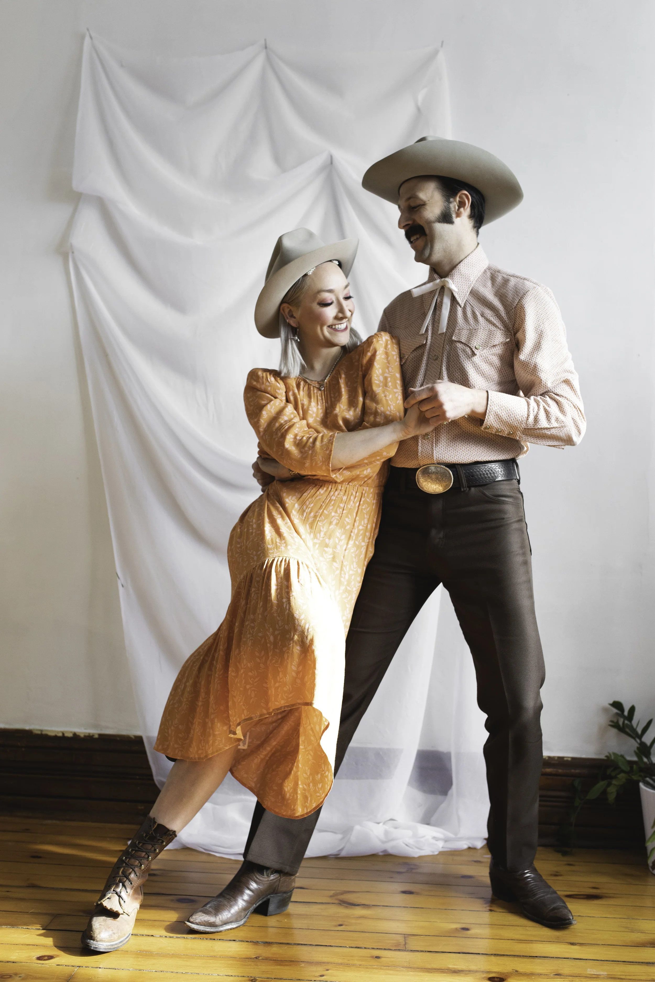 A man and woman dressed in cowboy attire dancing together on a wooden floor. The woman is wearing an orange dress and cowboy boots, while the man is wearing a pink shirt, brown pants, and cowboy boots. They are smiling and holding hands with a white fabric backdrop behind them.