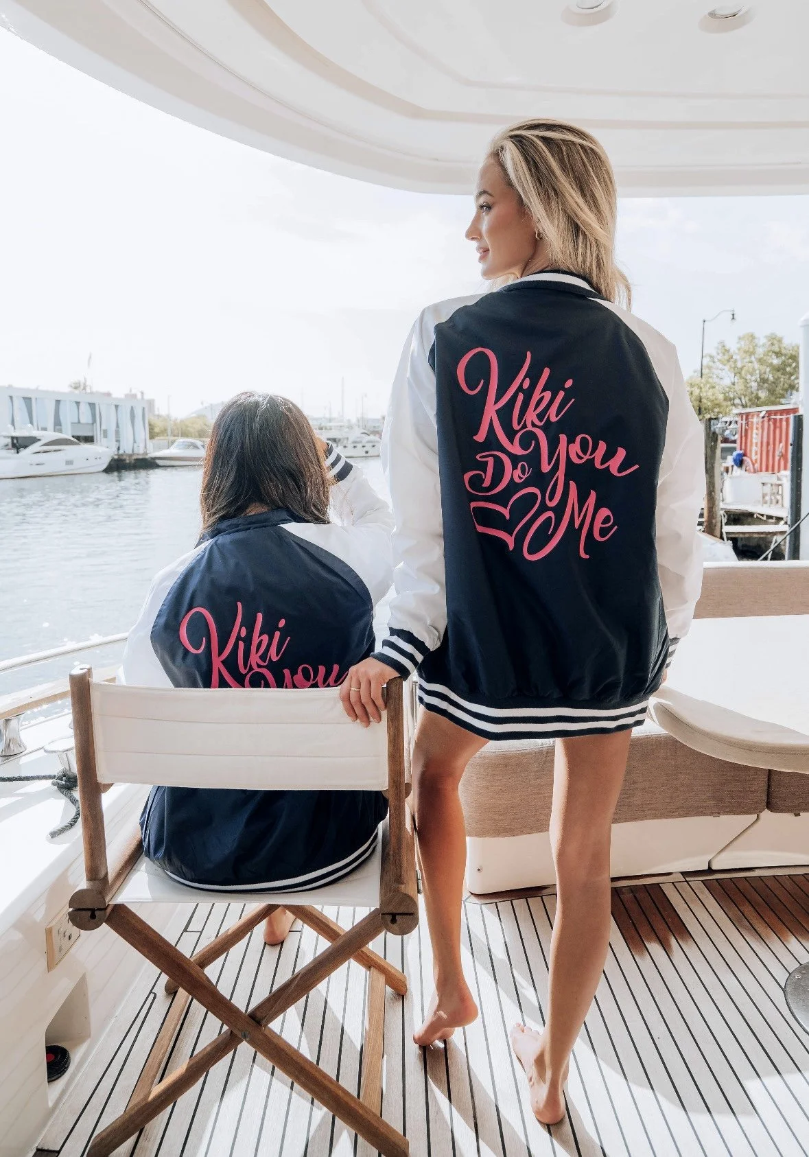 Two women wearing matching navy and white jackets with pink lettering, sitting and standing on a boat by the water. The jackets read 'Kiki Do You Love Me.' One woman is sitting on a chair, while the other is standing barefoot with one hand on a chair back, looking out at the water and boats in the background.