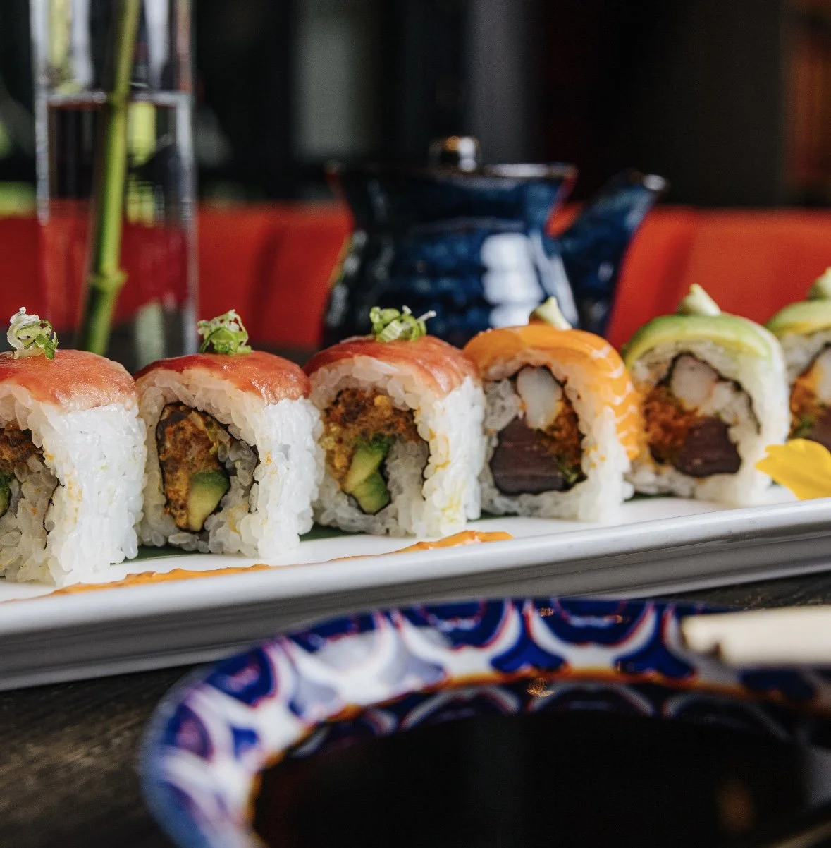 Close-up of assorted sushi rolls on a rectangular white plate, with soy sauce in a patterned bowl in the foreground and a blurred background of a restaurant setting.