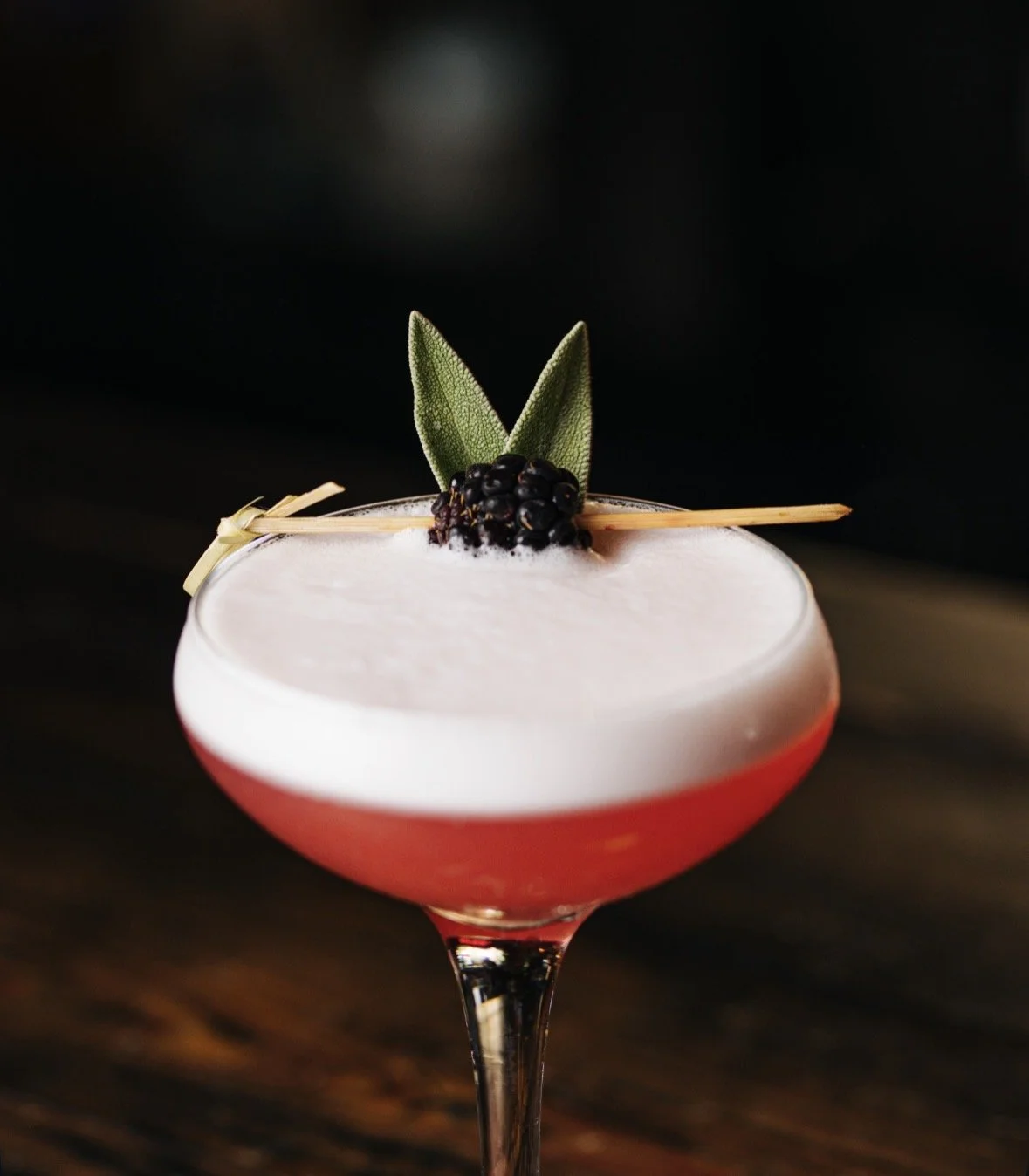 A cocktail with a pinkish-red bottom and a frothy white top, garnished with sage leaves, blackberries, and a decorative straw, served in a stemmed glass.