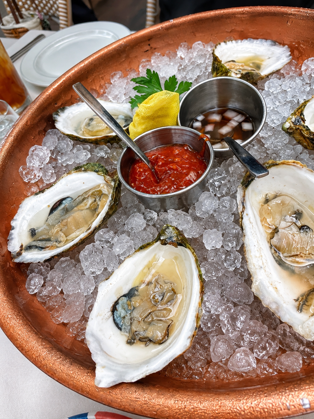 A copper bowl filled with ice and fresh oysters on the half shell, served with small containers of cocktail sauce and mignonette, garnished with lemon, parsley, and a yellow cloth.
