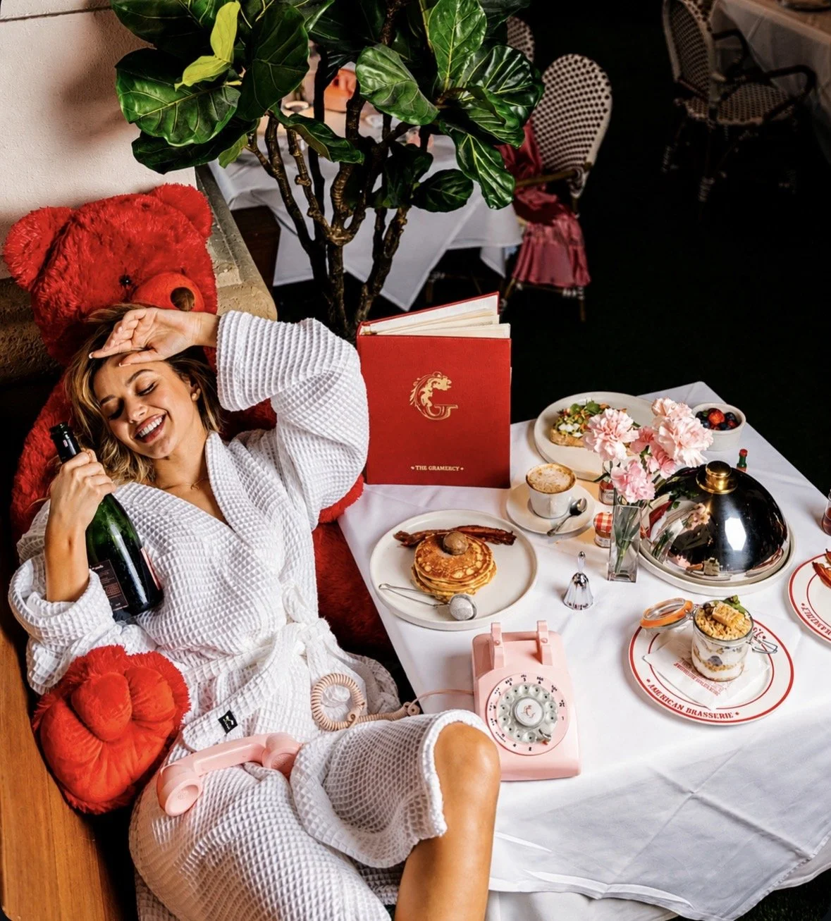 A woman in a white bathrobe reclining at a table filled with food and drinks, smiling and holding a bottle of wine. The table has pancakes, a flower arrangement, a vintage telephone, and various desserts. She appears relaxed and happy.