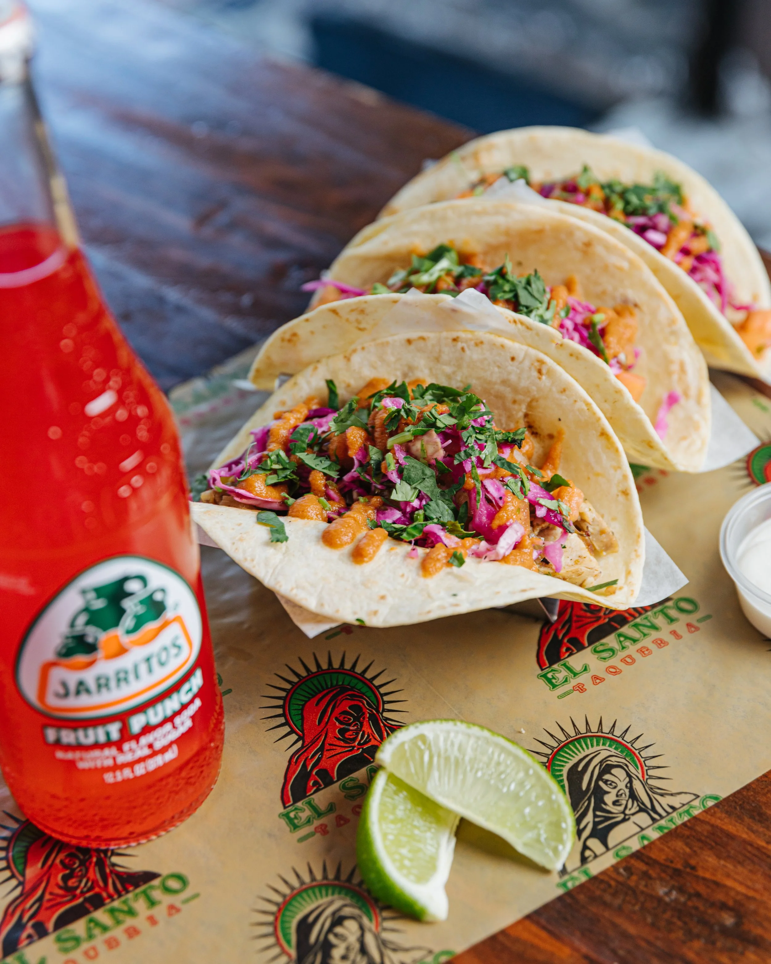 Three tacos filled with shredded meat, chopped cilantro, and pink cabbage served on El Sinaloense restaurant branded paper, with a lime wedge and a bottle of hot sauce on a wooden table.