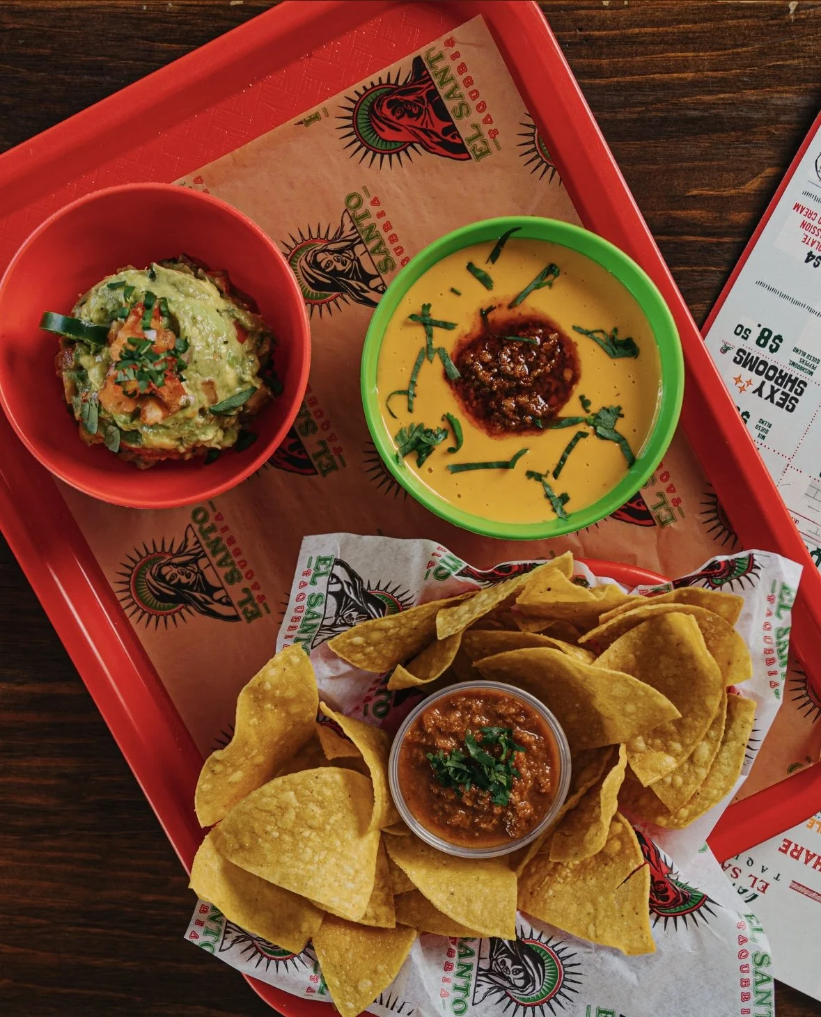 A tray of Mexican food including tortilla chips with salsa, guacamole, and yellow sauce with chili, on a paper liner with restaurant logo, and a drink menu on the side.