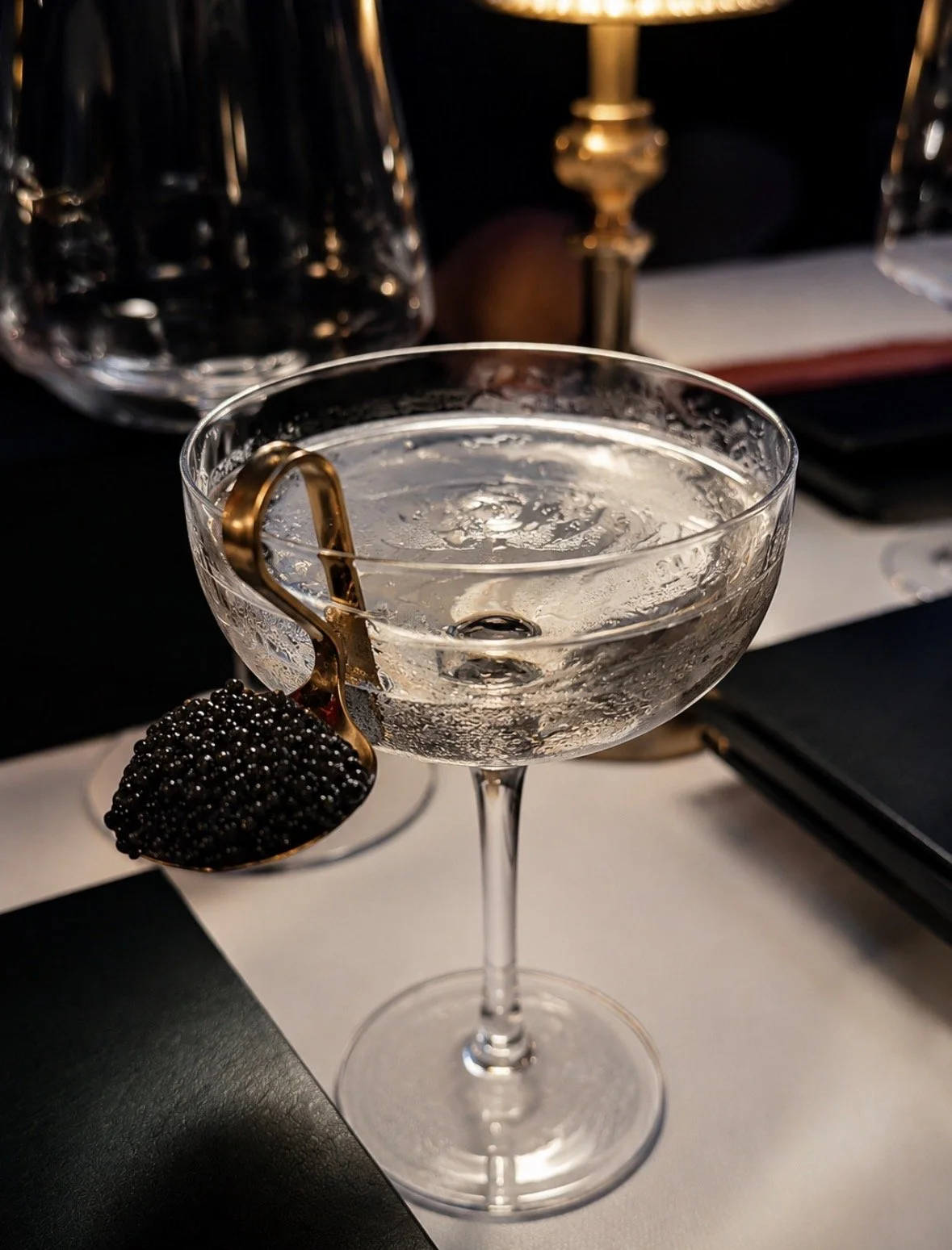 A cocktail glass filled with a clear beverage, garnished with a gold-colored or gold-accented ring holding a black caviar or caviar-like garnish, on a table with dark and light elements, in a sophisticated setting.