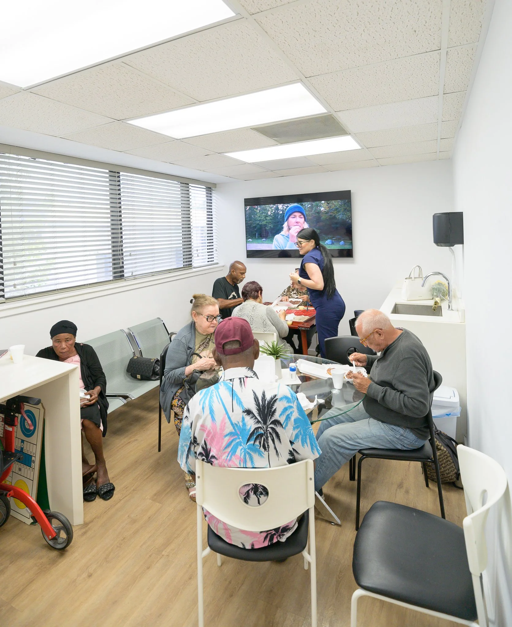 People sitting and socializing in a bright, white room with blinds, with some eating and others conversing around tables.