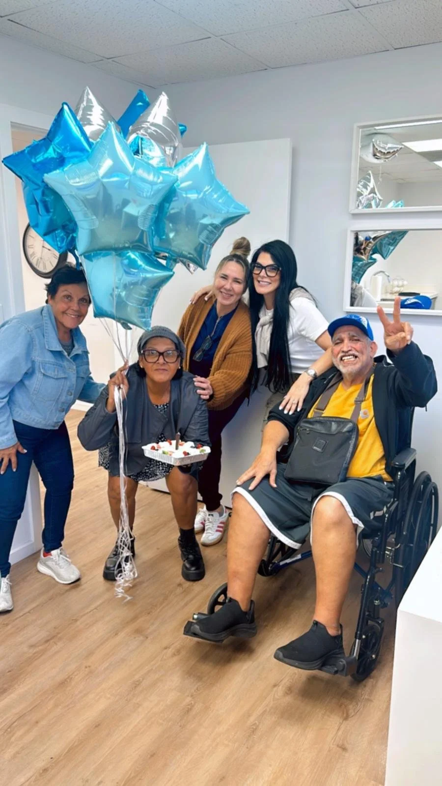 Group of five people celebrating with balloons and a cake in a room, including a person in a wheelchair.