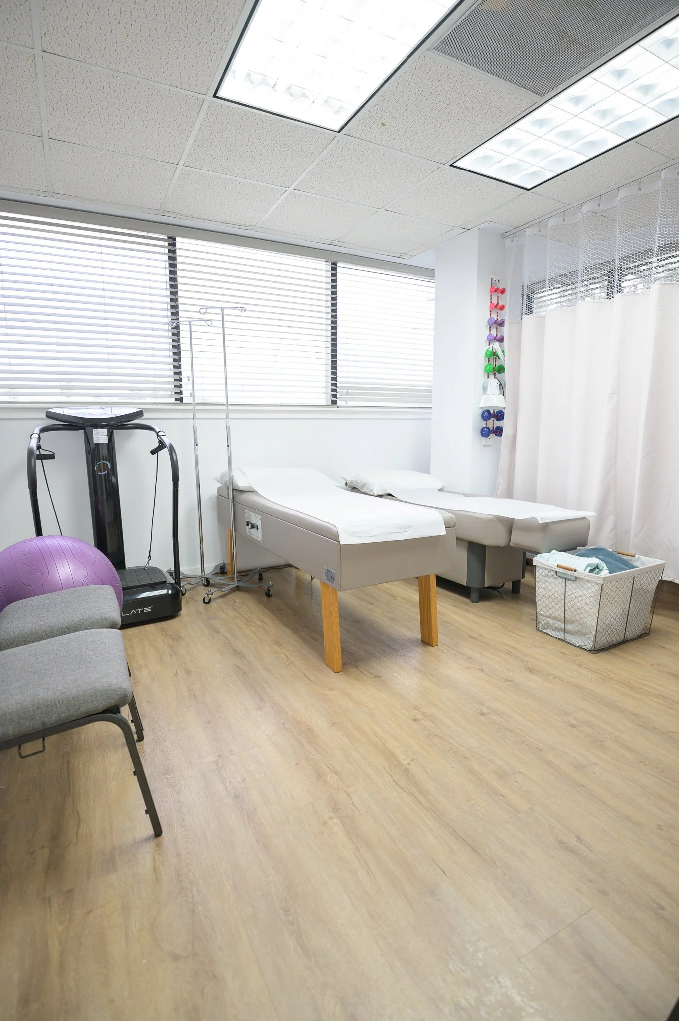 A medical examination room with a hospital bed, medical equipment, a chair, a purple exercise ball, and large windows with blinds, illuminated by ceiling lights.