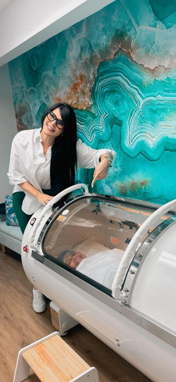 A woman with long black hair, glasses, and a white shirt is smiling and pointing at a baby in a hospital bassinet. The baby is lying down, covered with a white blanket. The background features a colorful, abstract blue and brown mural.