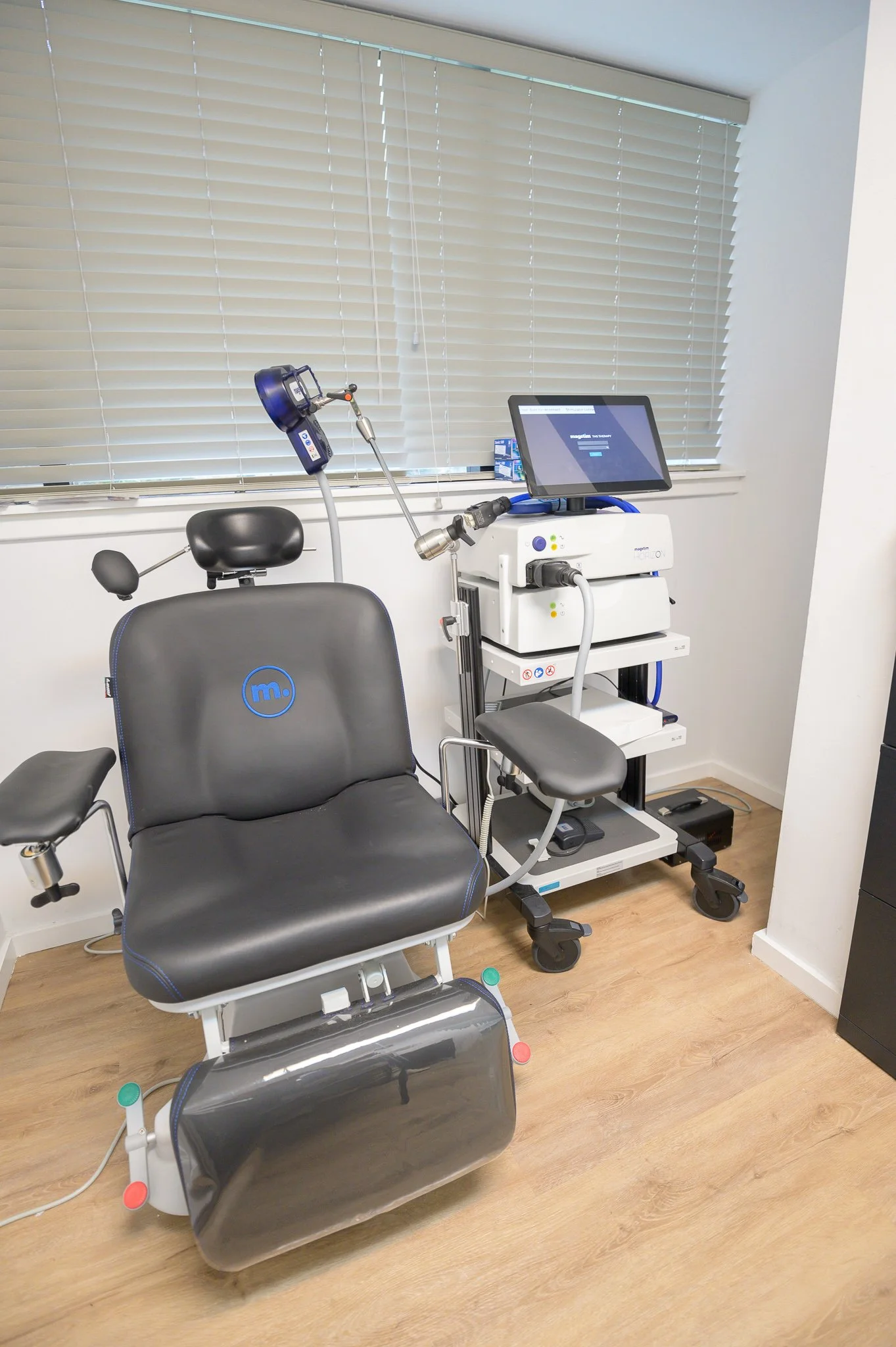 Medical office with a black examination chair and medical equipment, including a monitor and diagnostic tools, near a window with closed blinds.