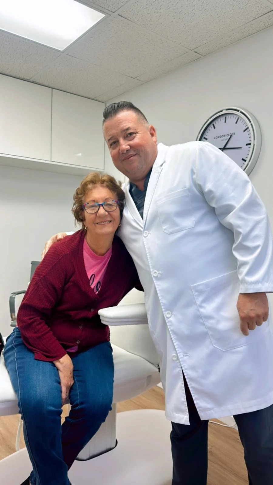 A woman and a man in a medical office, smiling and posing for the camera. The woman is seated on an examination table, wearing glasses, a pink shirt, and a red cardigan. The man, likely a doctor, is standing next to her, dressed in a white lab coat. There is a clock on the wall behind them showing the time as 2:10.