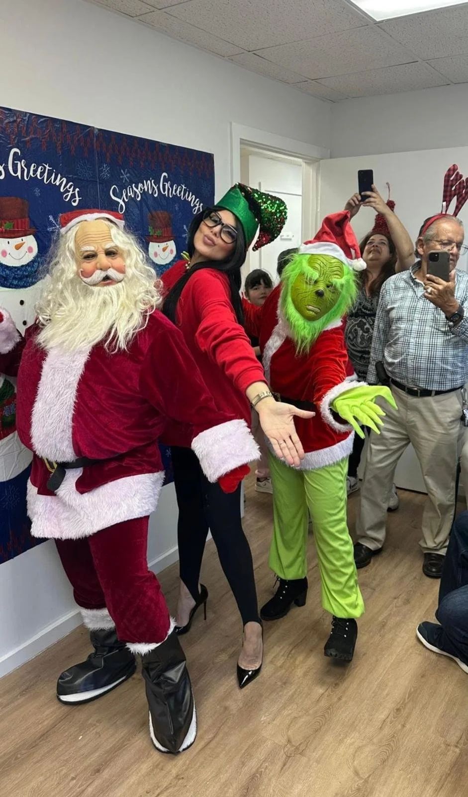 People dressed in Christmas costumes, including Santa Claus and a Grinch, taking a group photo at a holiday party.